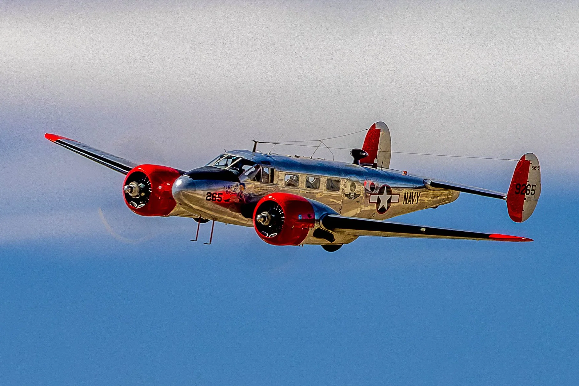 A vintage U.S. Navy aircraft flying in the sky with polished metal body and red accents on engines and tail.