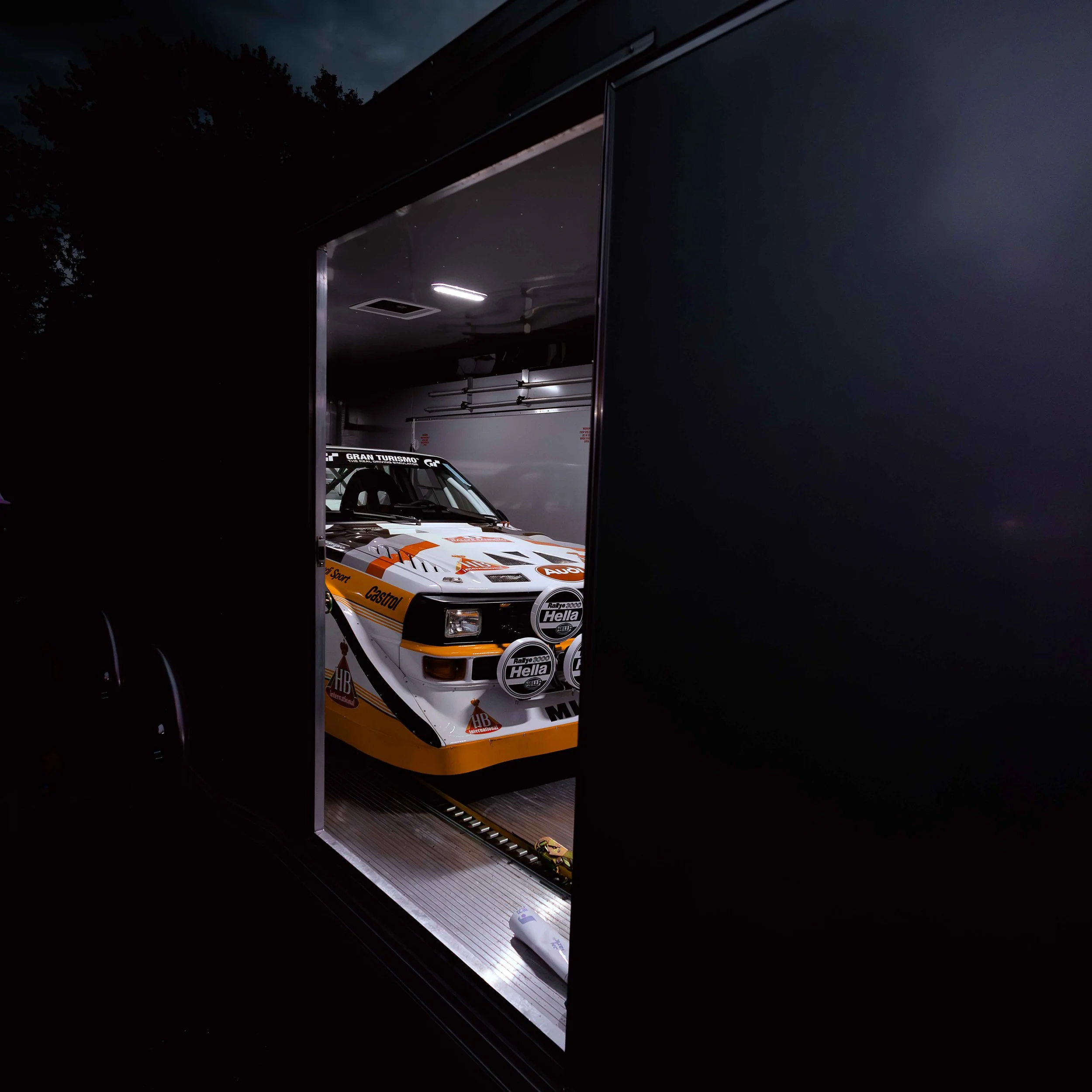 A vintage race car inside a dark trailer with some interior lights on, showing the front end with sponsors' logos and the rally car's orange and white livery.