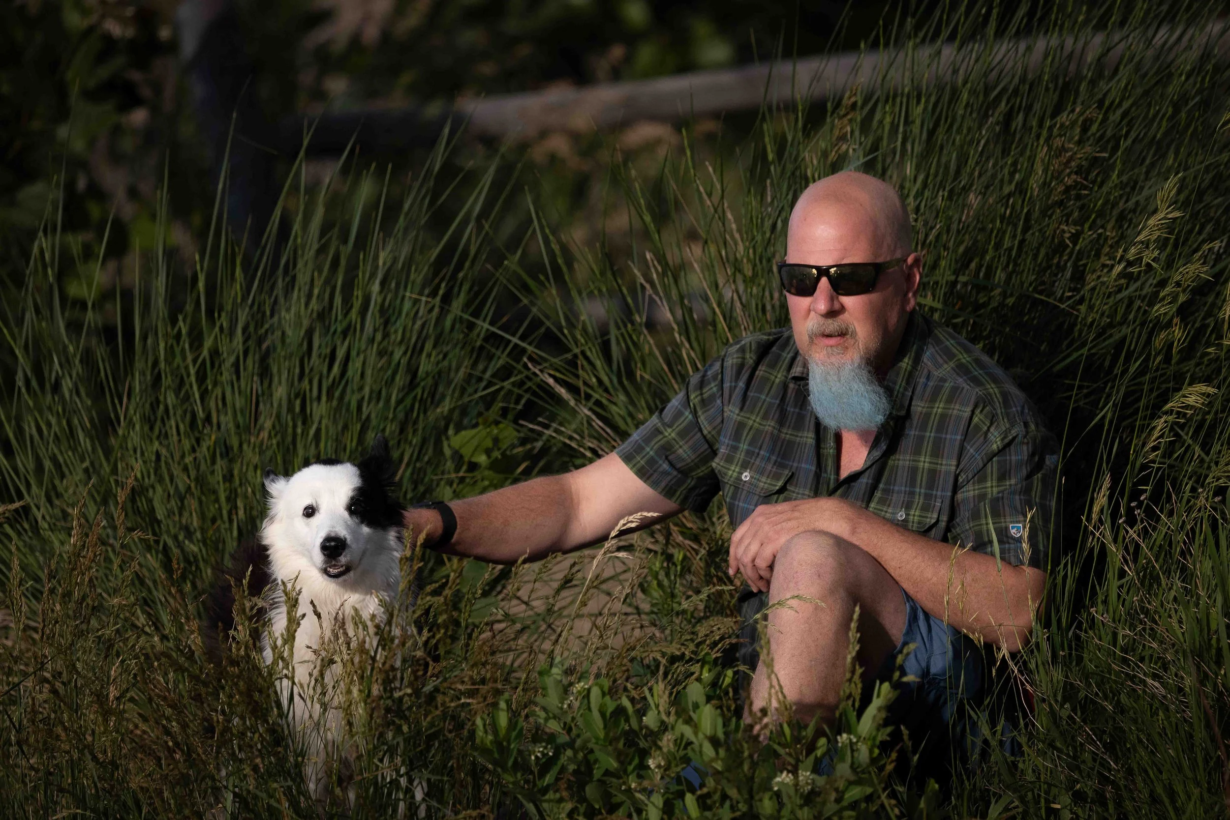 A man with sunglasses and a blue beard kneeling in grassy outdoors with a black and white dog.