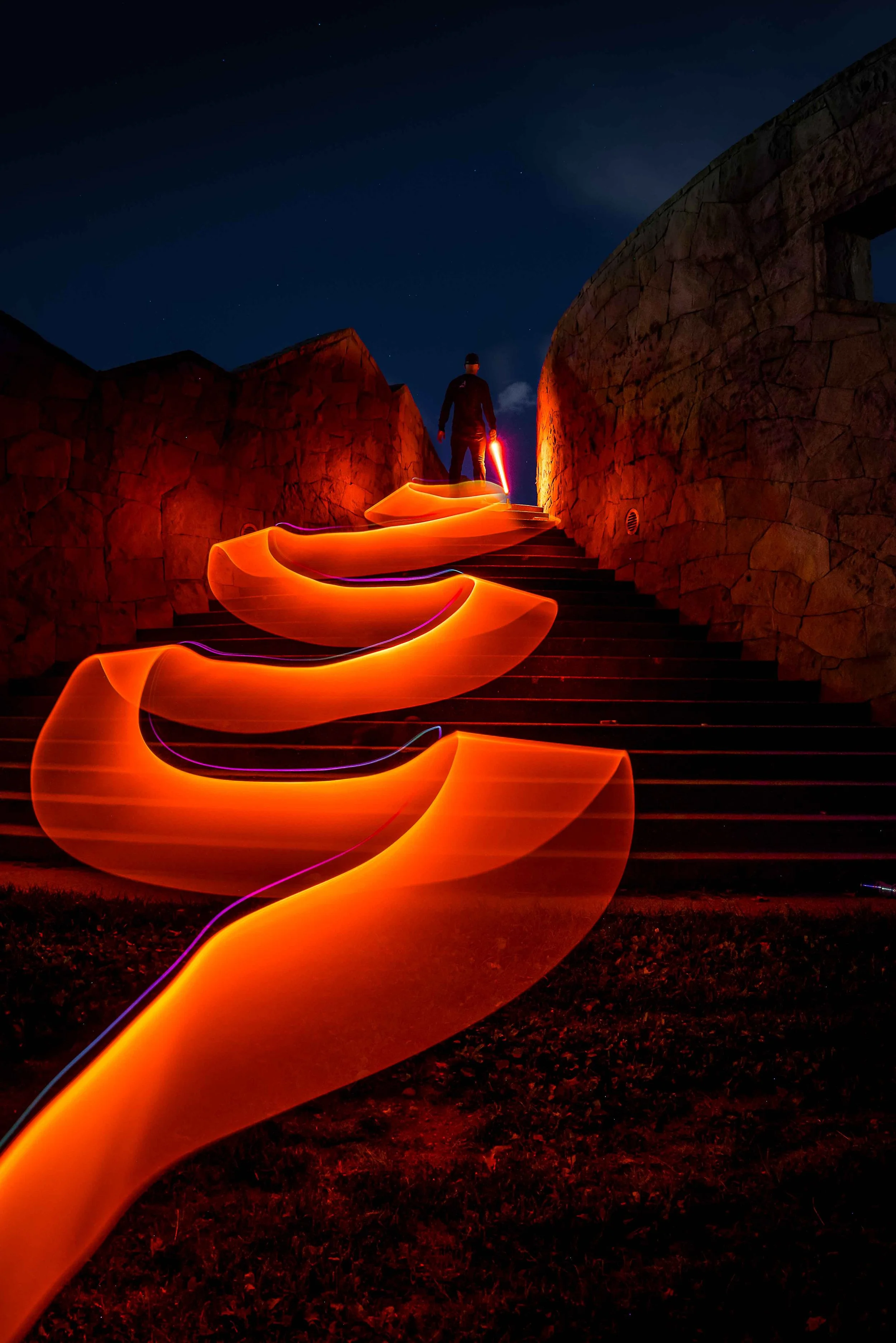A person stands on stairs at night holding a glowing red light, with swirling orange light trails in front of them and a starry sky above.