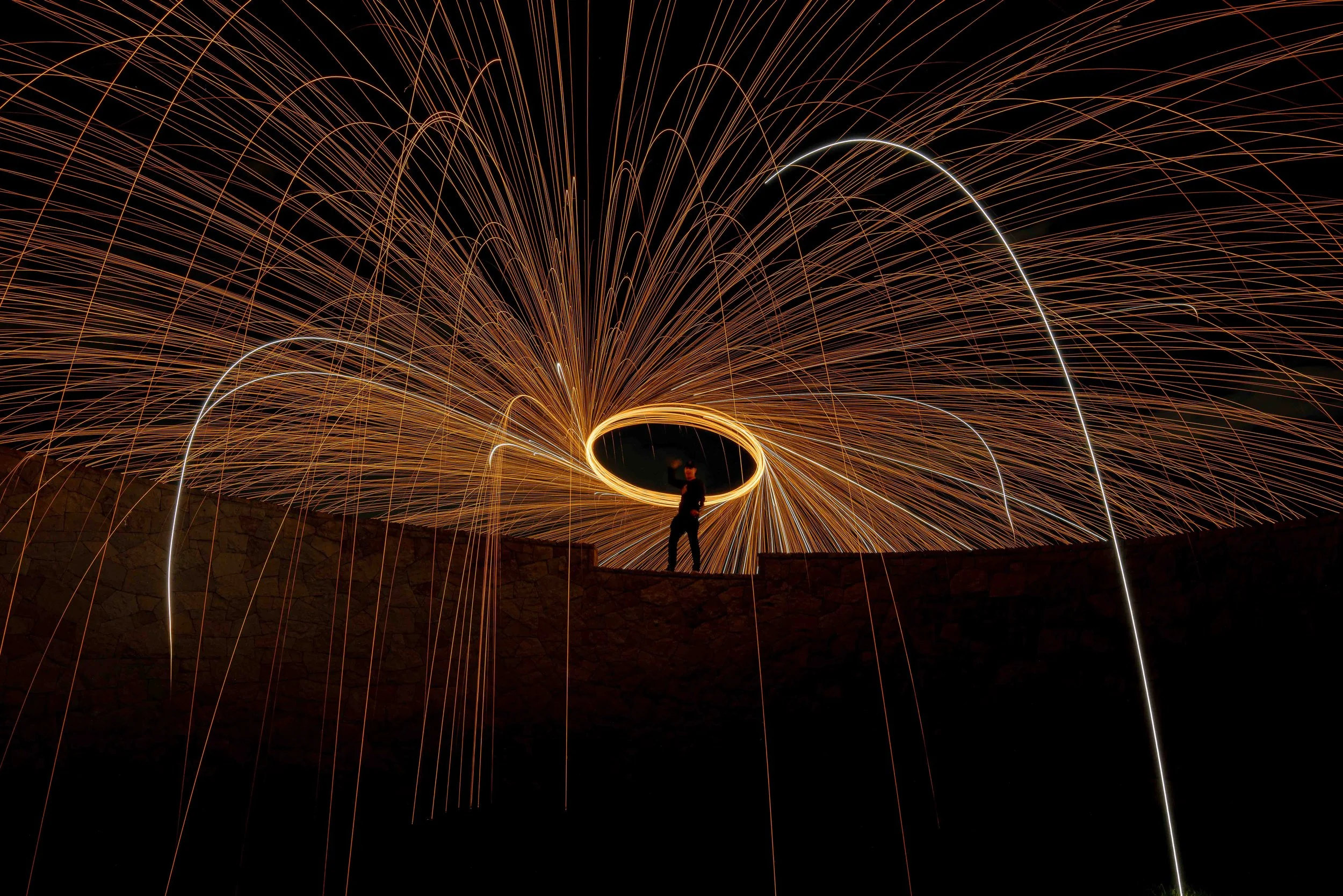 Person standing on a ledge with long exposure light streaks and sparks creating circular patterns around them at night.