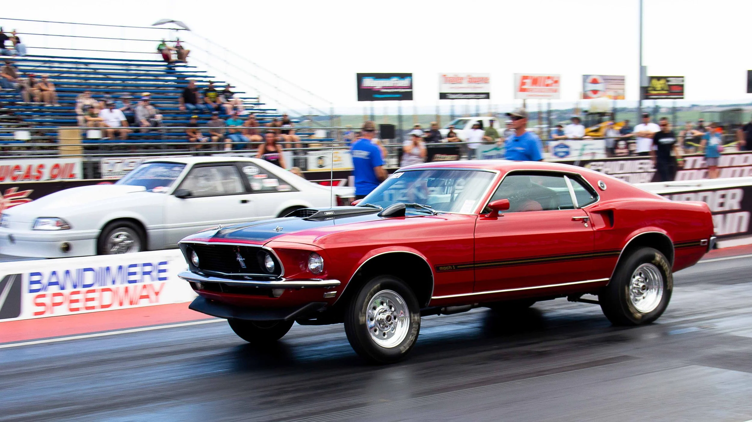 A red classic Ford Mustang Mach 1 race car on a drag strip with a black stripe on its hood, wheels lifted off the ground as it accelerates, with a white car behind, spectators in the stands, and banners reading 'BANDIMERE SPEEDWAY' in the background.