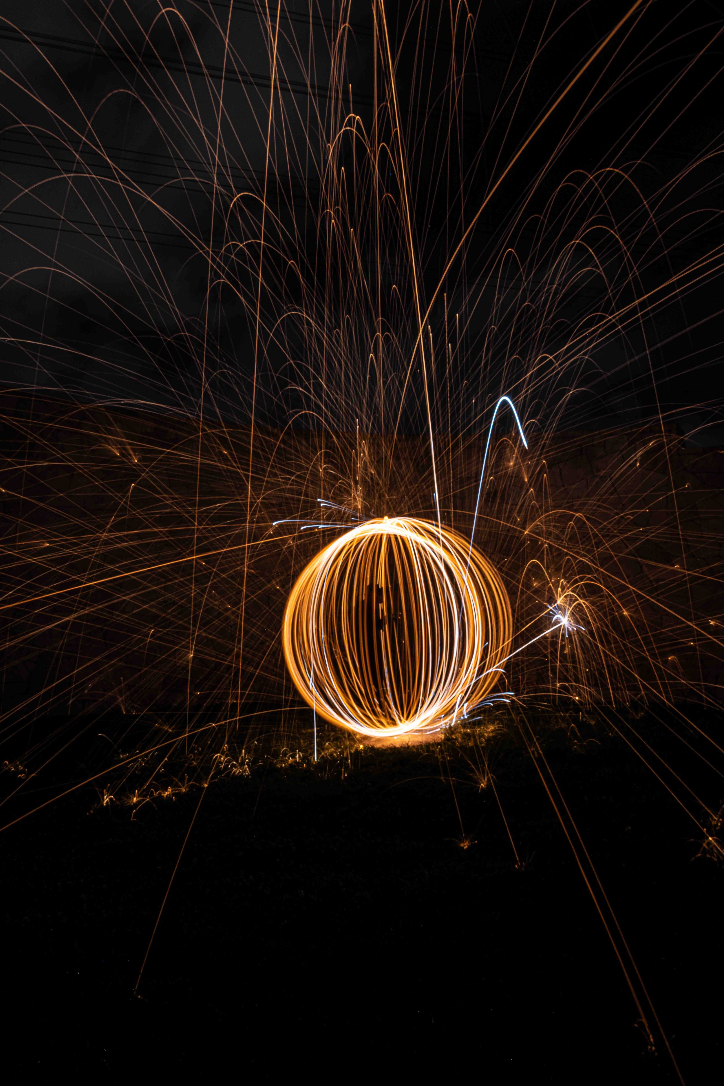 A long exposure photograph of steel wool spinning creates a sphere with bright sparks flying outward against a dark background.