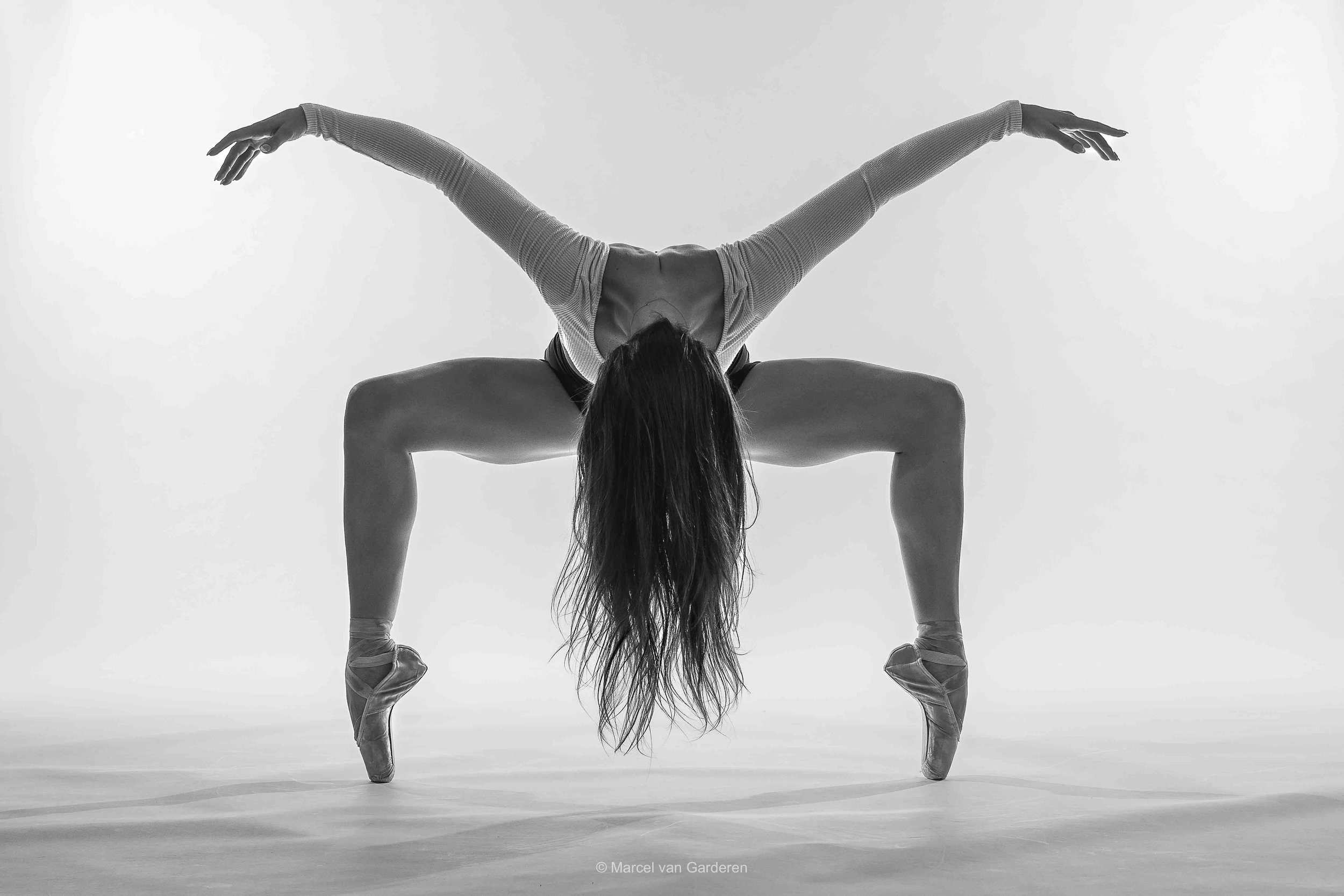 A dancer performs a pose with her hands on the ground and legs bent, wearing pointe shoes, in a high-contrast black and white photo.