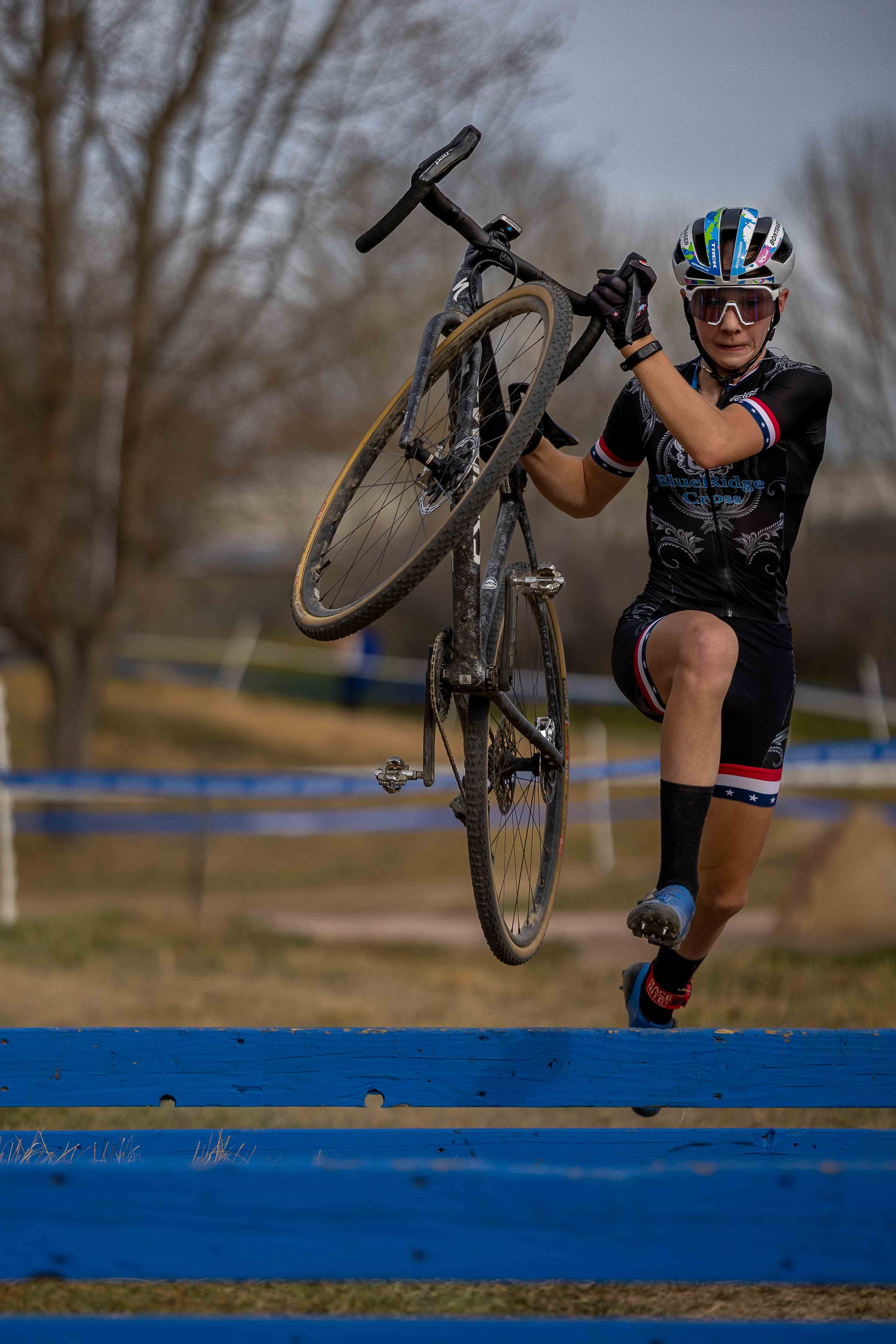A cyclist in a black uniform and helmet jumping over a blue barrier on a cross-country course while holding a mountain bike.