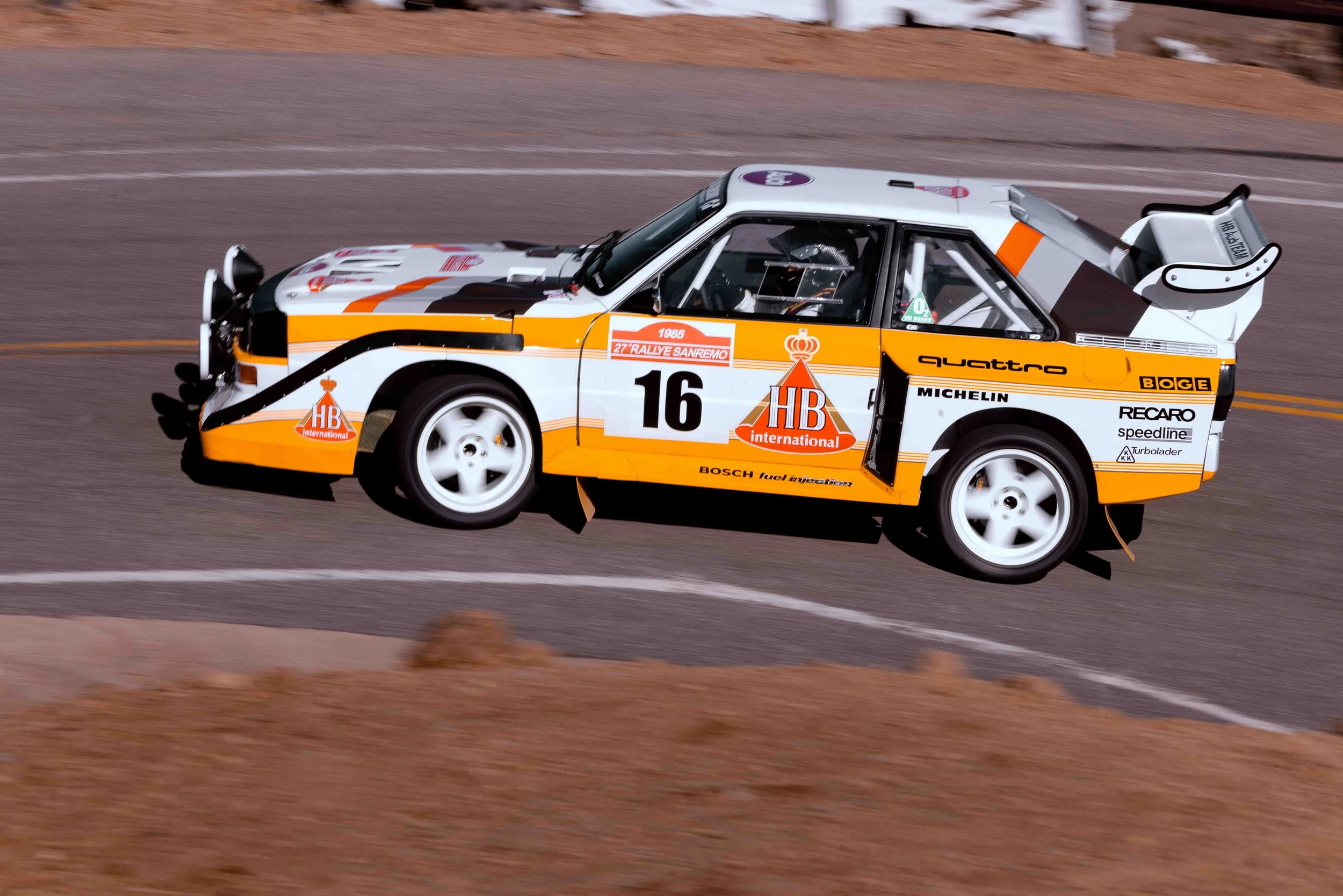 A vintage race car, primarily in white with orange and black accents, speeding on a road with desert landscape in the background. The car has racing decals and the number 16 on its side.