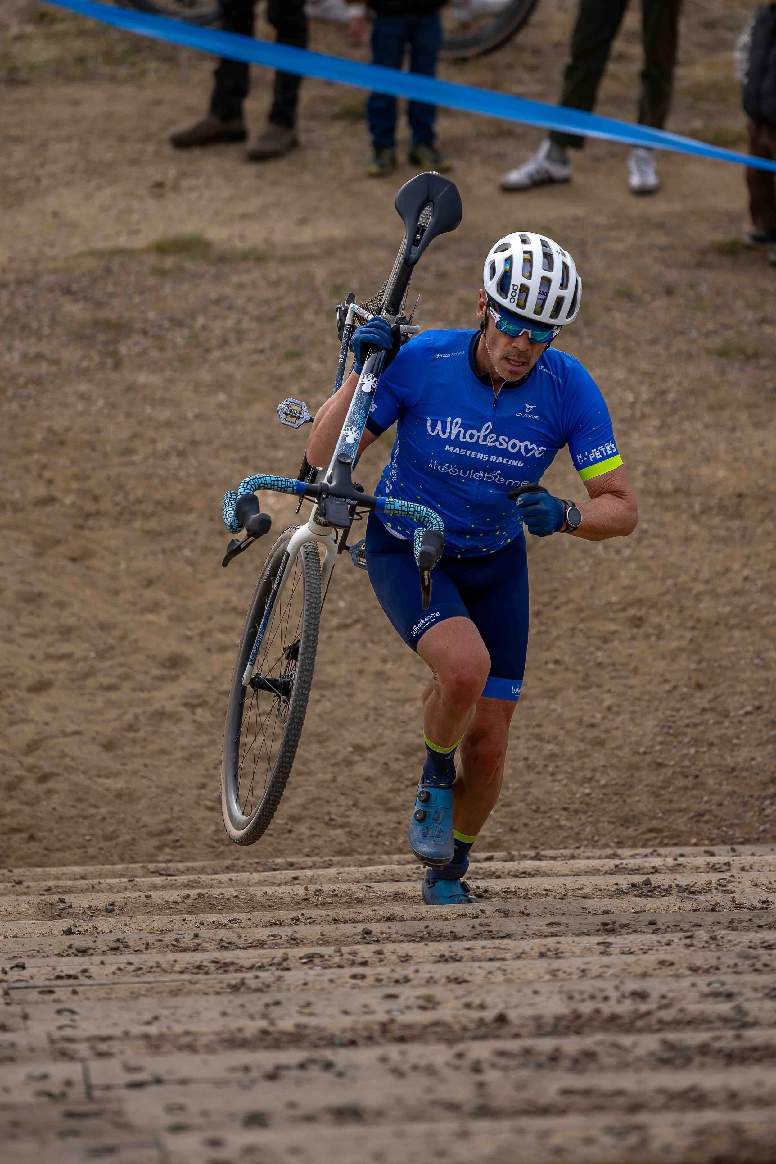 A male cyclist in blue gear carrying a bicycle on his shoulder during a race on a dirt trail.