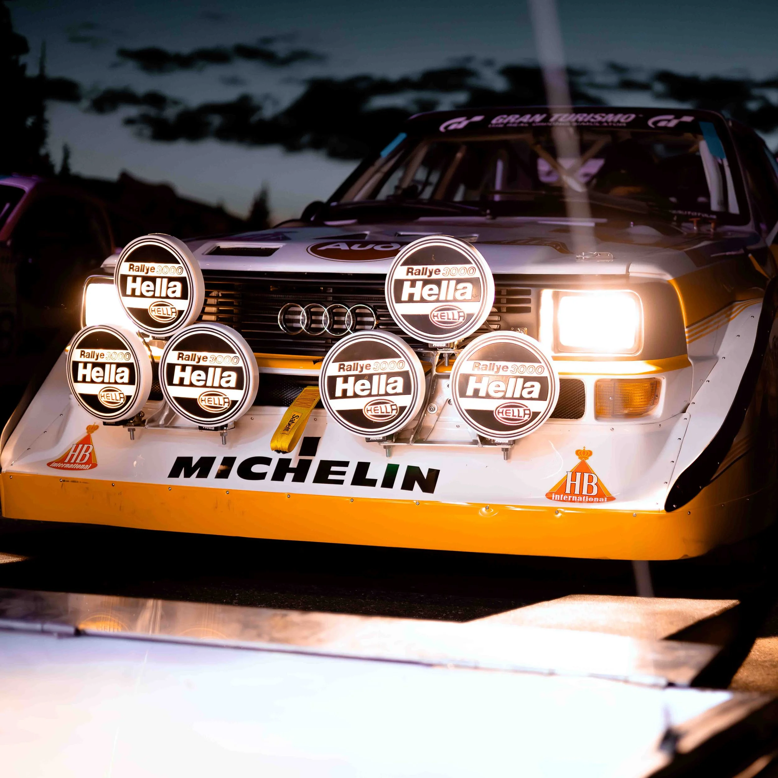 A vintage rally car at dusk with four large rally lights mounted on the front, displaying 'Rallye 3000 Hella' logos, and sponsorship logos for 'Michelin' and 'HB International'.