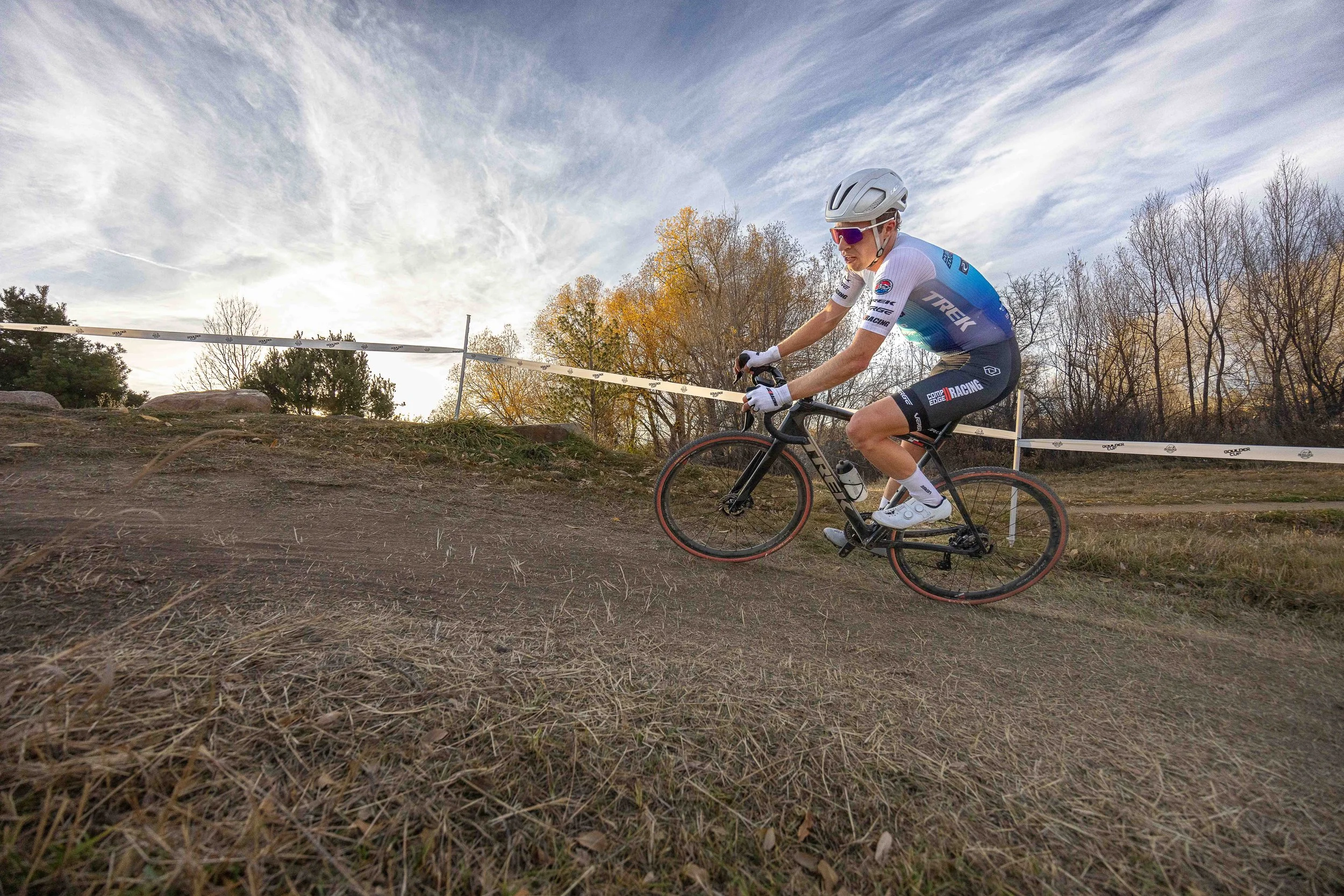 Cyclist wearing a helmet and team jersey riding a mountain bike on a dirt trail during a race, with trees and a partly cloudy sky in the background.