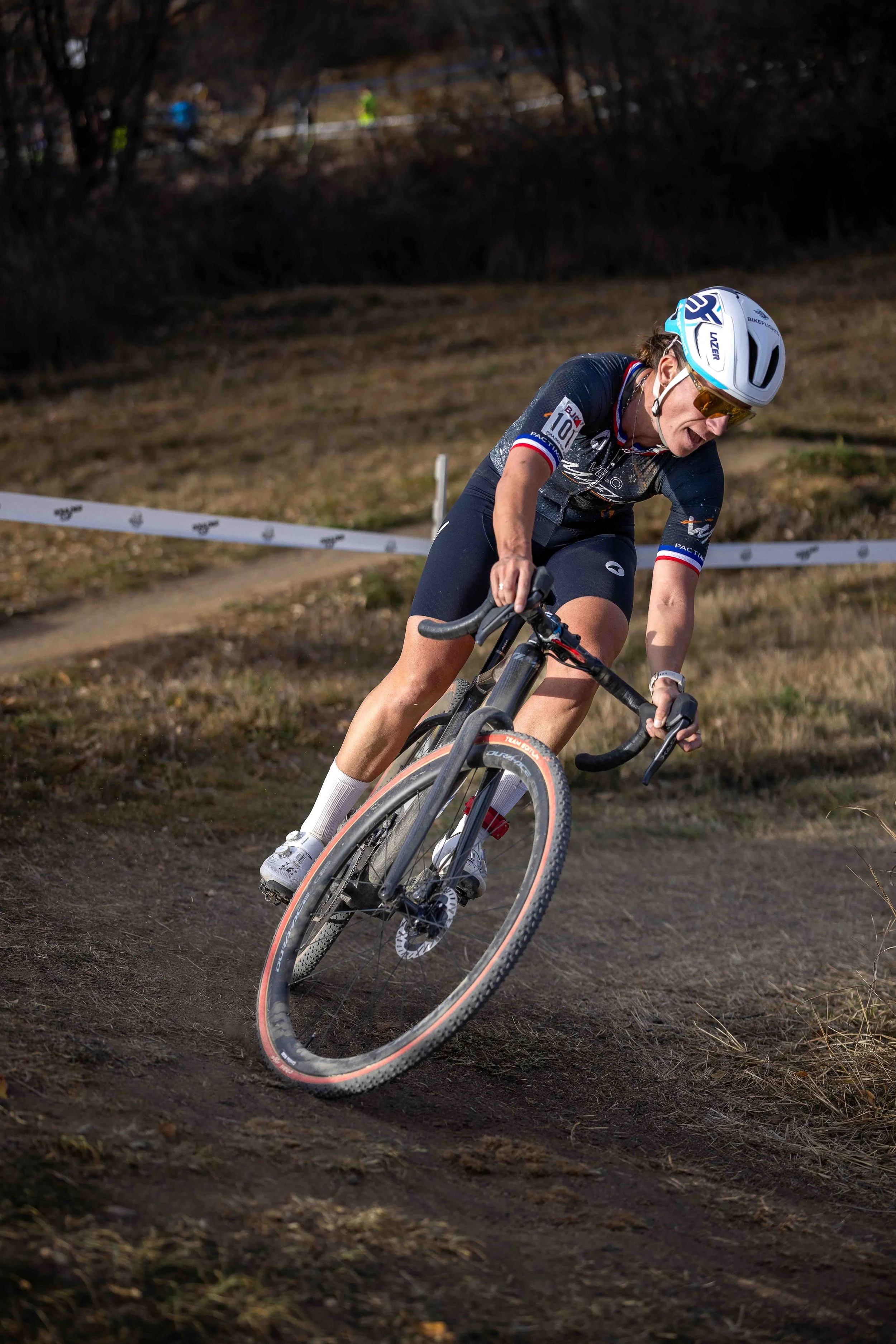 A female cyclist racing on a dirt course, wearing a white helmet, sunglasses, and athletic gear, leaning into a turn.