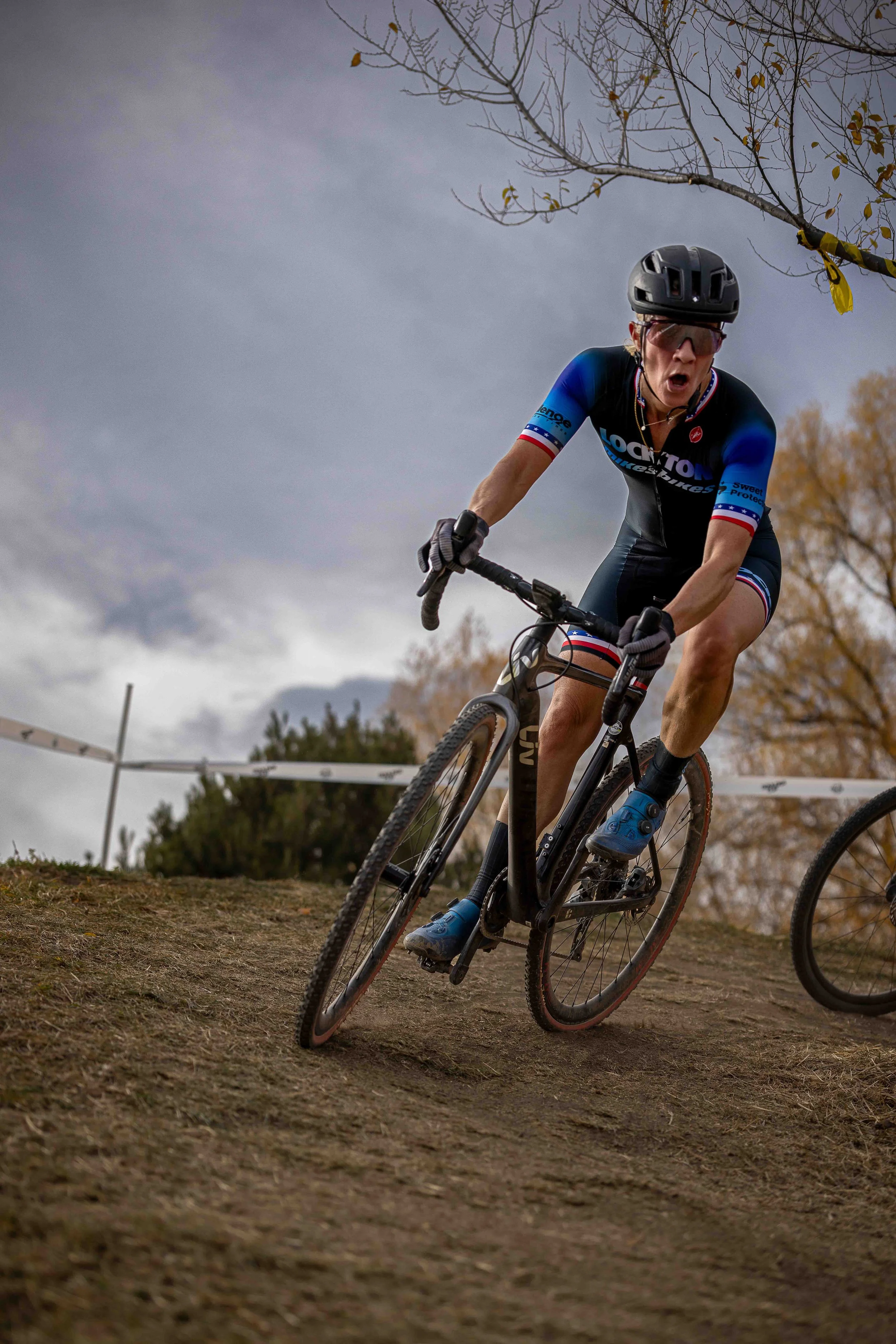 A woman wearing a black helmet and cycling gear is riding a mountain bike on a dirt trail, leaning into a turn with a focused expression on her face, under a cloudy sky with some trees in the background.