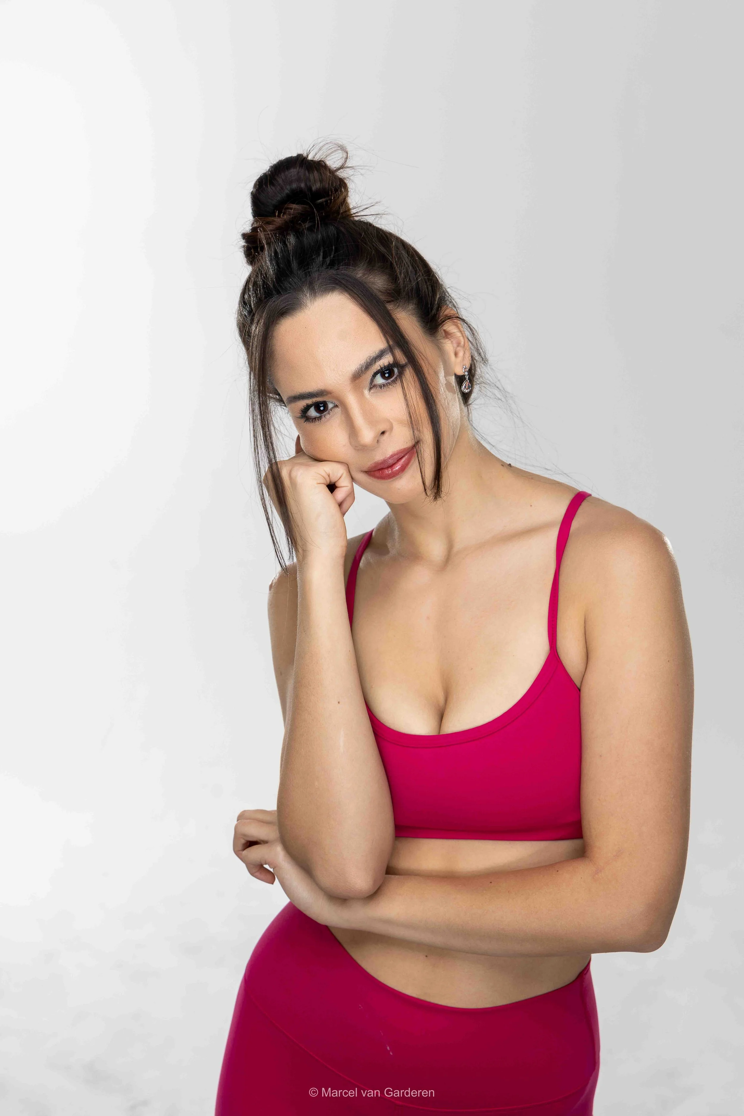 A woman with dark hair styled in a messy bun wearing a pink sports bra and matching leggings, looking at the camera with her chin resting on her hand.