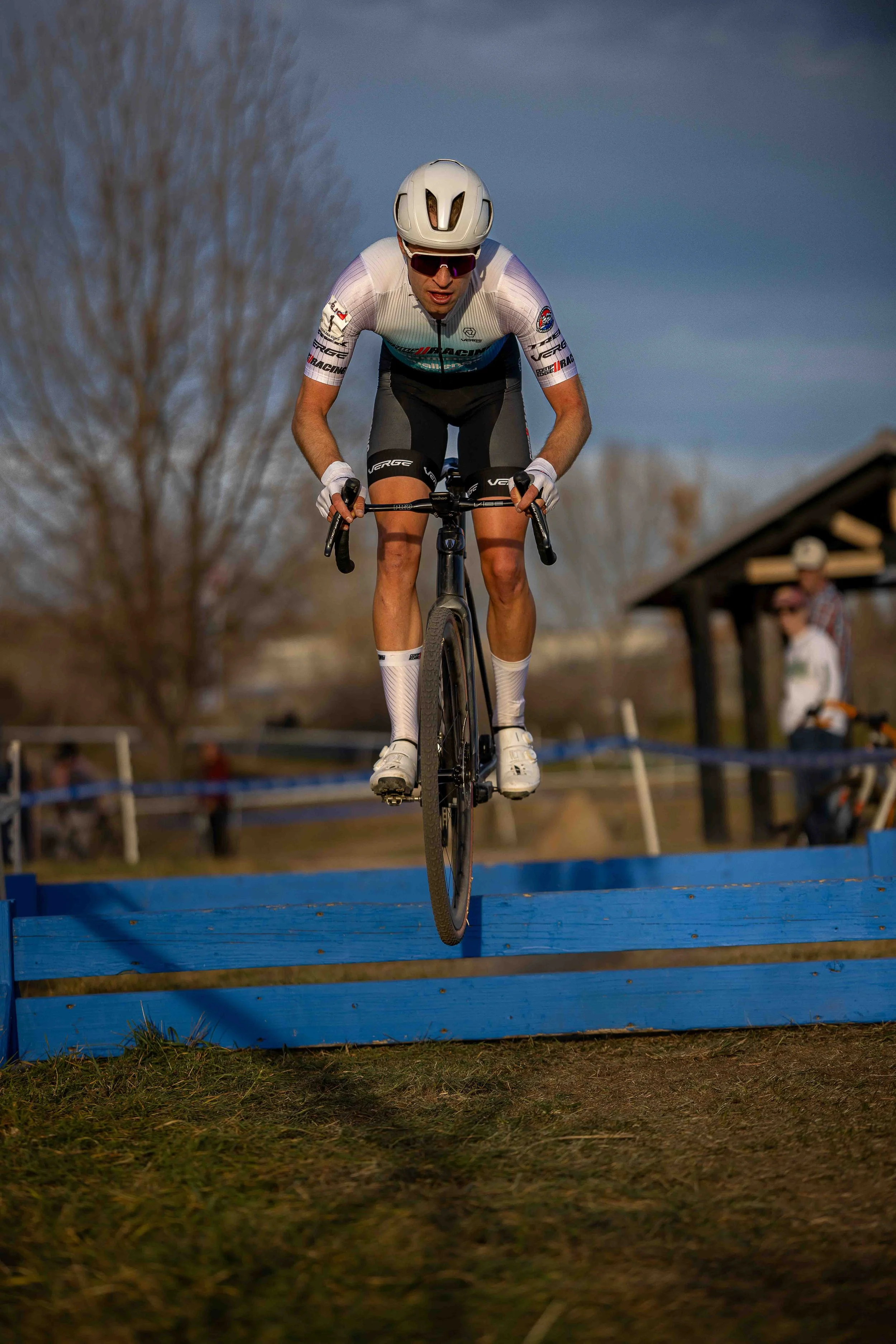 A cyclist wearing a white helmet, sunglasses, and a white cycling jersey is jumping over a blue obstacle on a bike trail outdoors, with trees and people in the background.