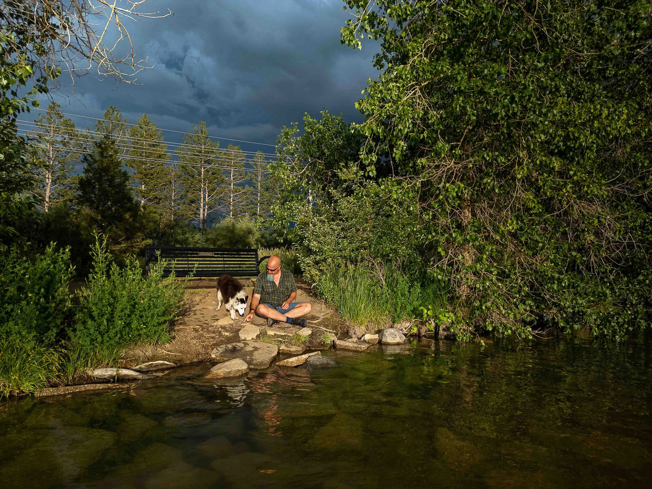 A man with a dog sitting on rocks by a riverbank with lush green trees and a cloudy sky in the background.