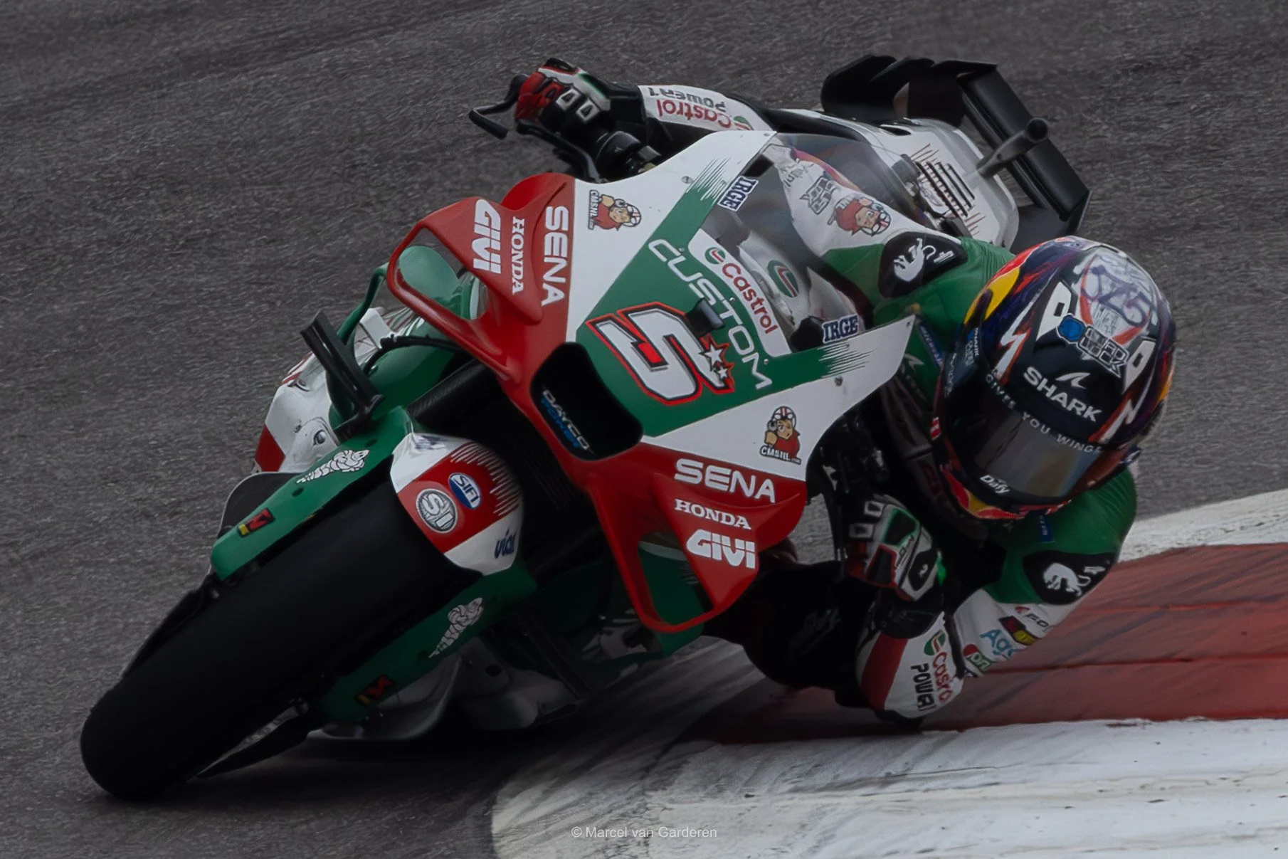 A motorcycle racer leaning into a turn on a racetrack, riding a bike with red, white, and green racing colors and various sponsor logos.