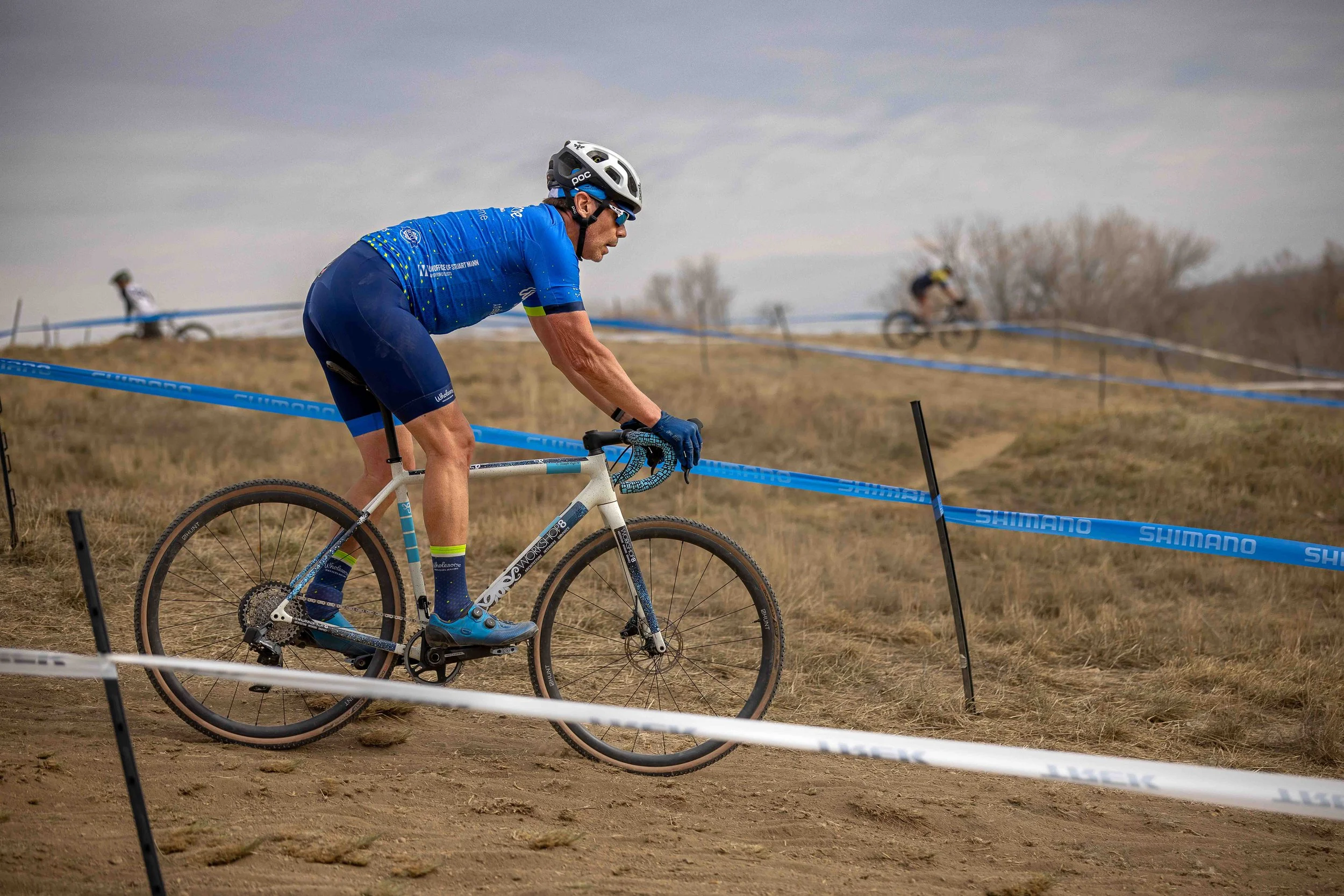 A male cyclist in a blue outfit riding a mountain bike on a dirt trail with a racing event marked by blue and white safety tape.