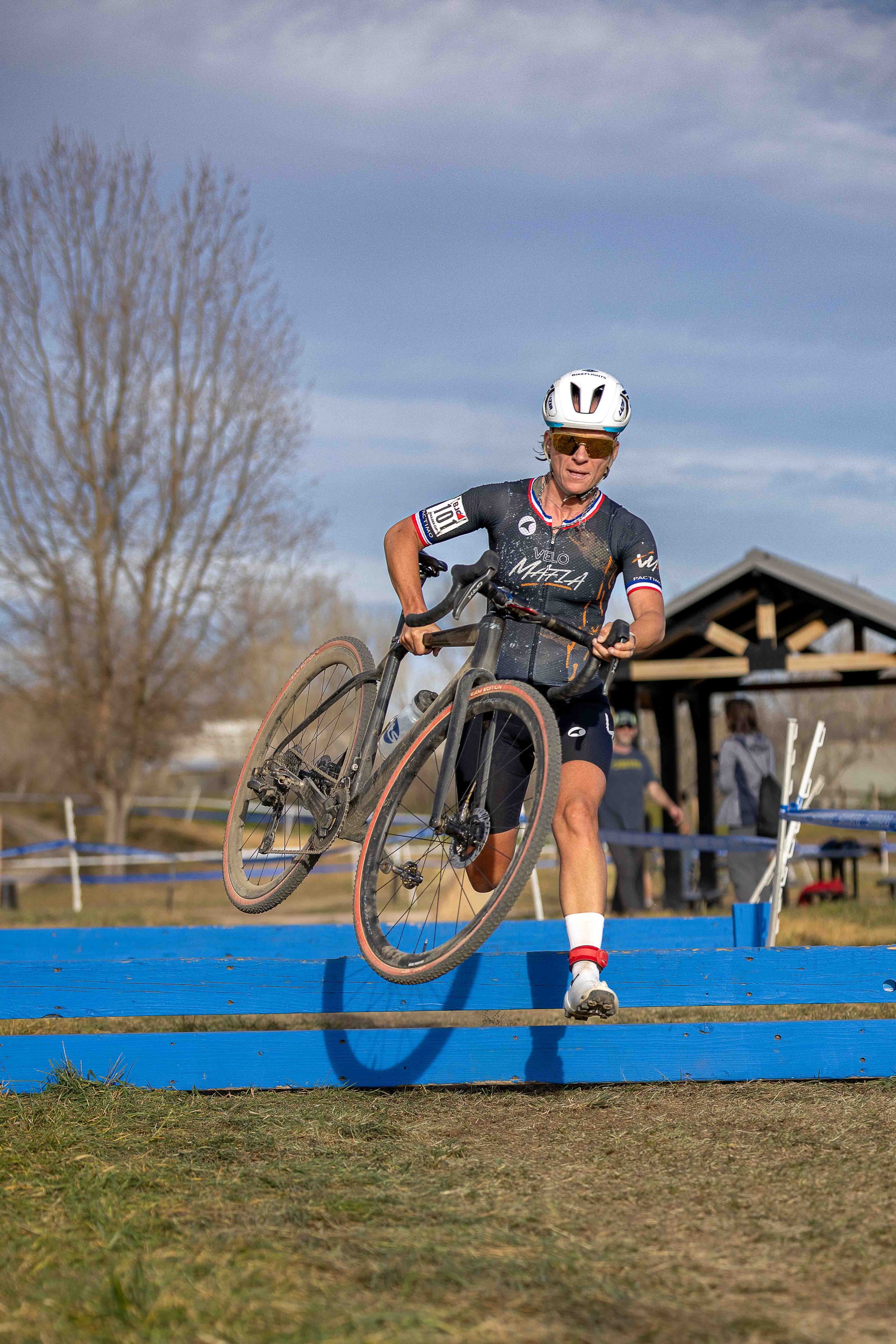 A cyclist in athletic gear jumping over a blue obstacle during a race, with trees and a pavilion in the background.