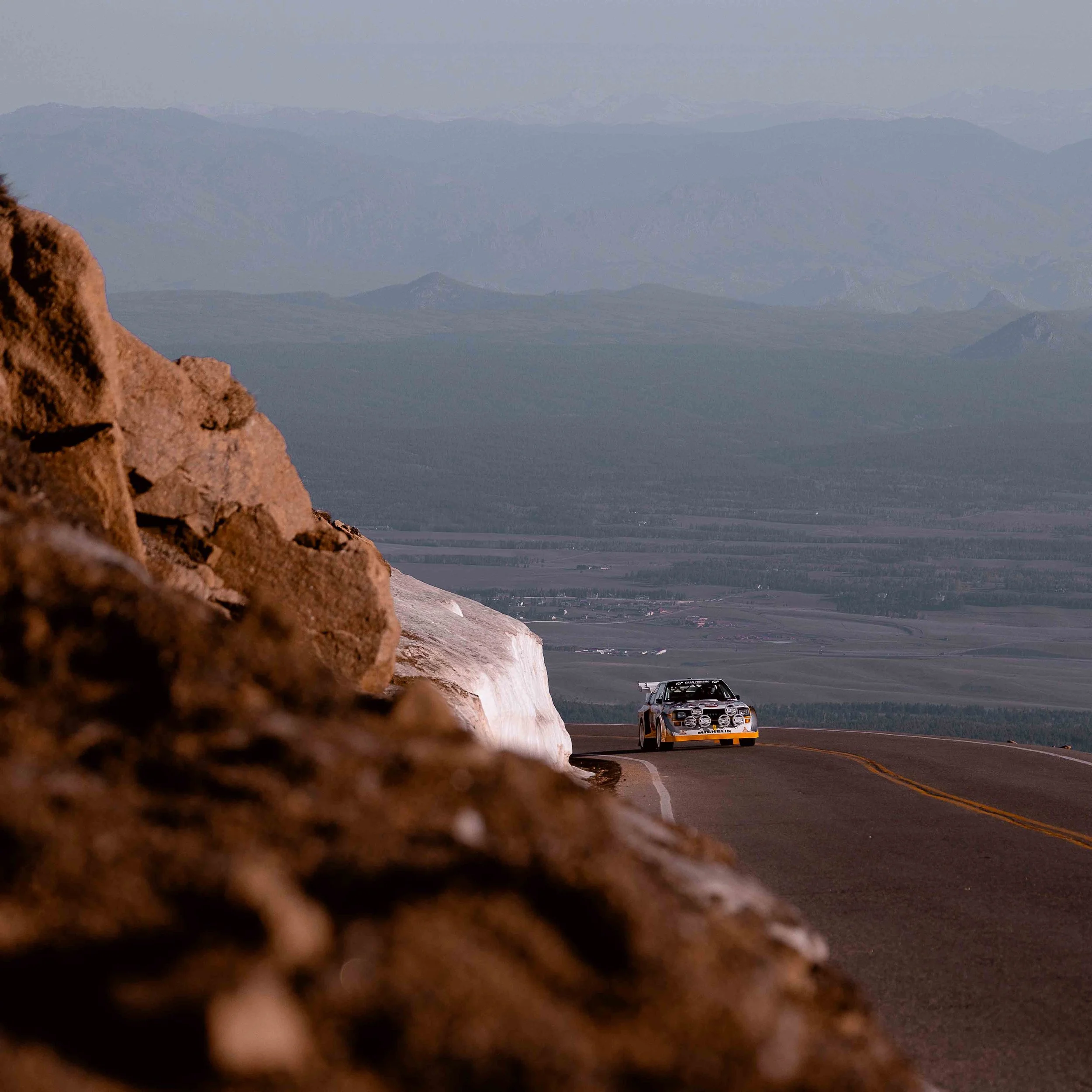 A white race car with yellow accents driving on a winding mountain road, with a rocky terrain in the foreground and a vast valley with distant mountains in the background.