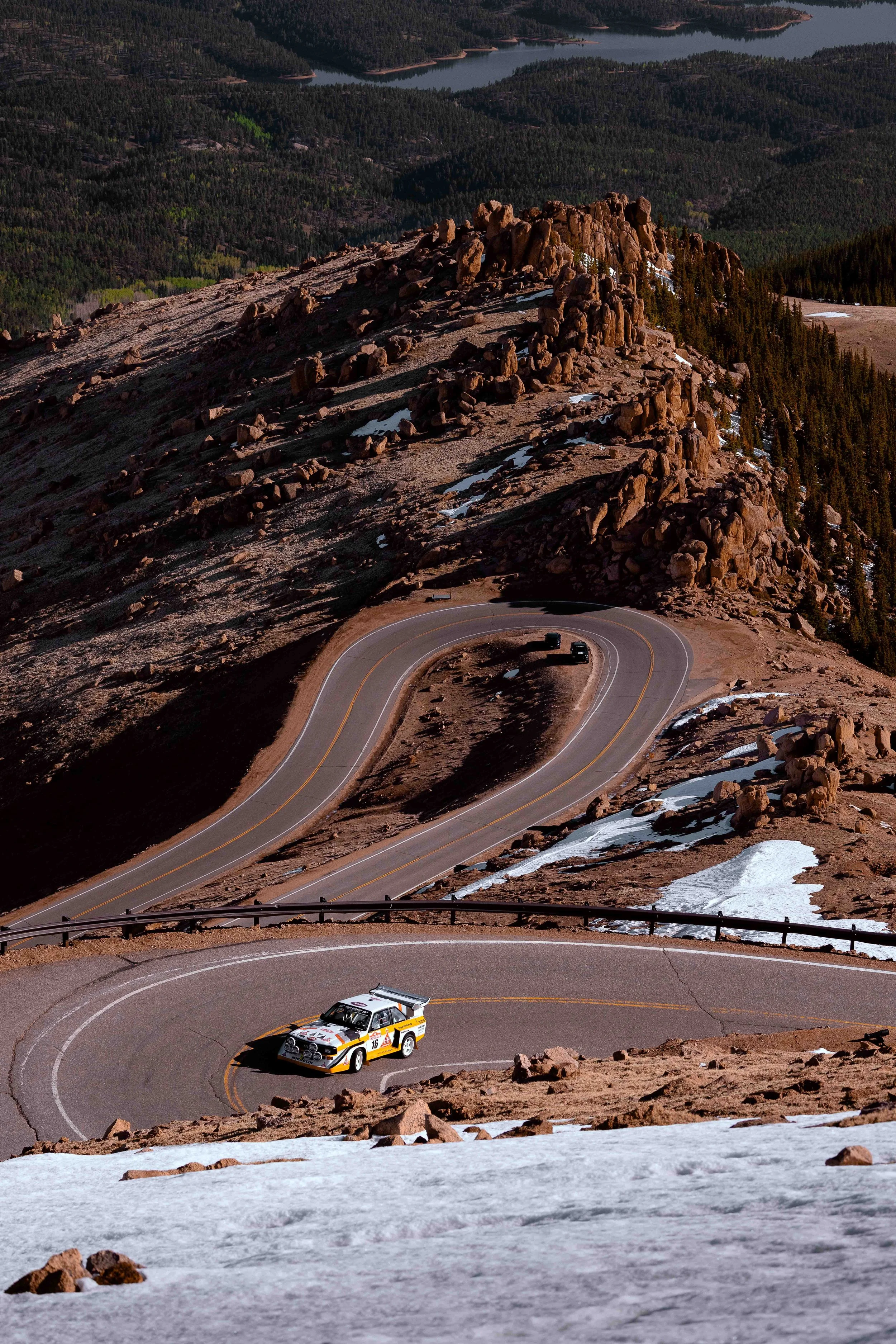 A race car on a winding mountain road with snowy patches, rocky terrain, and forested hills in the background.