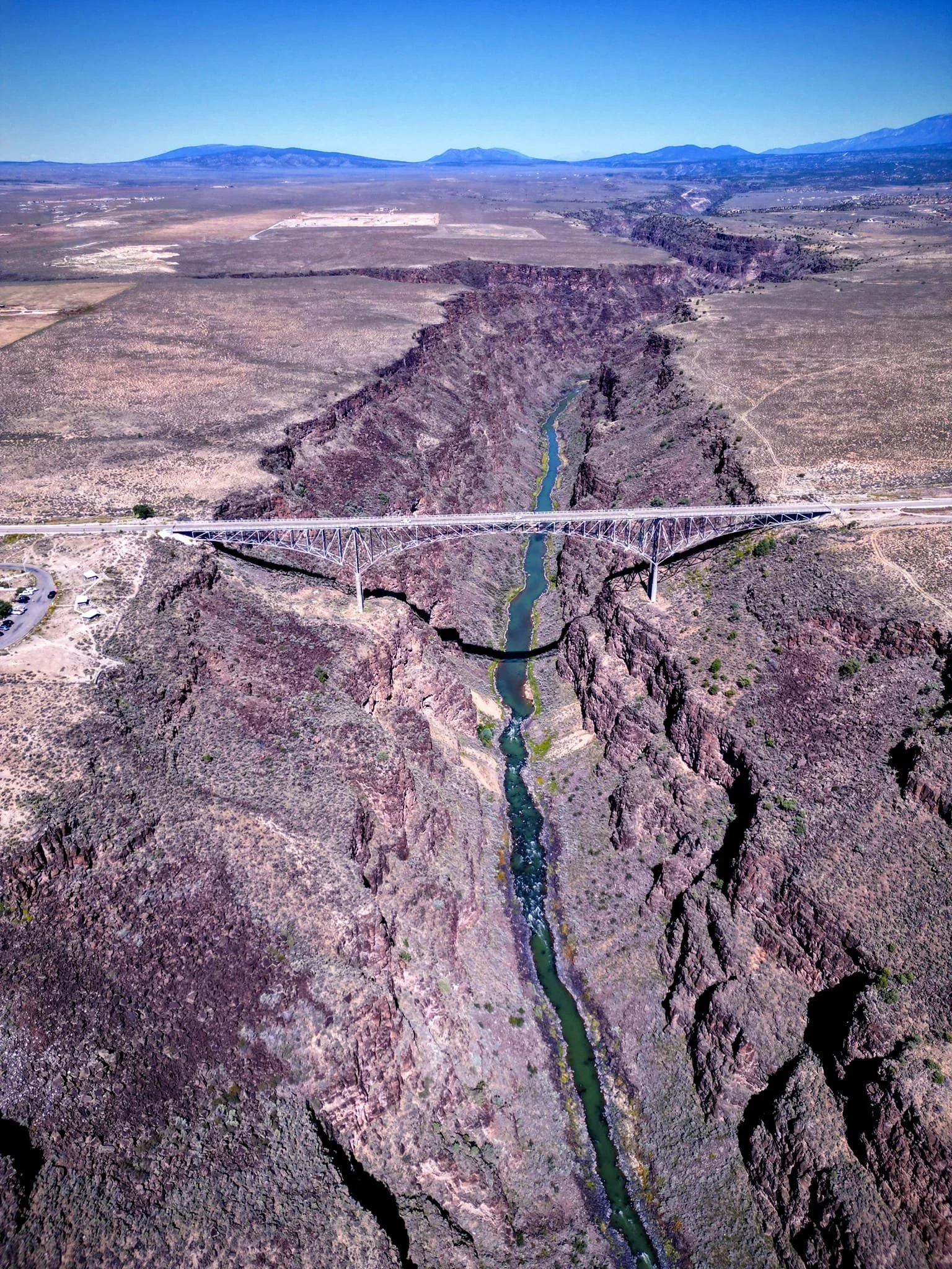 Aerial view of a bridge spanning a canyon with a river running through it, surrounded by desert landscape and distant mountains.