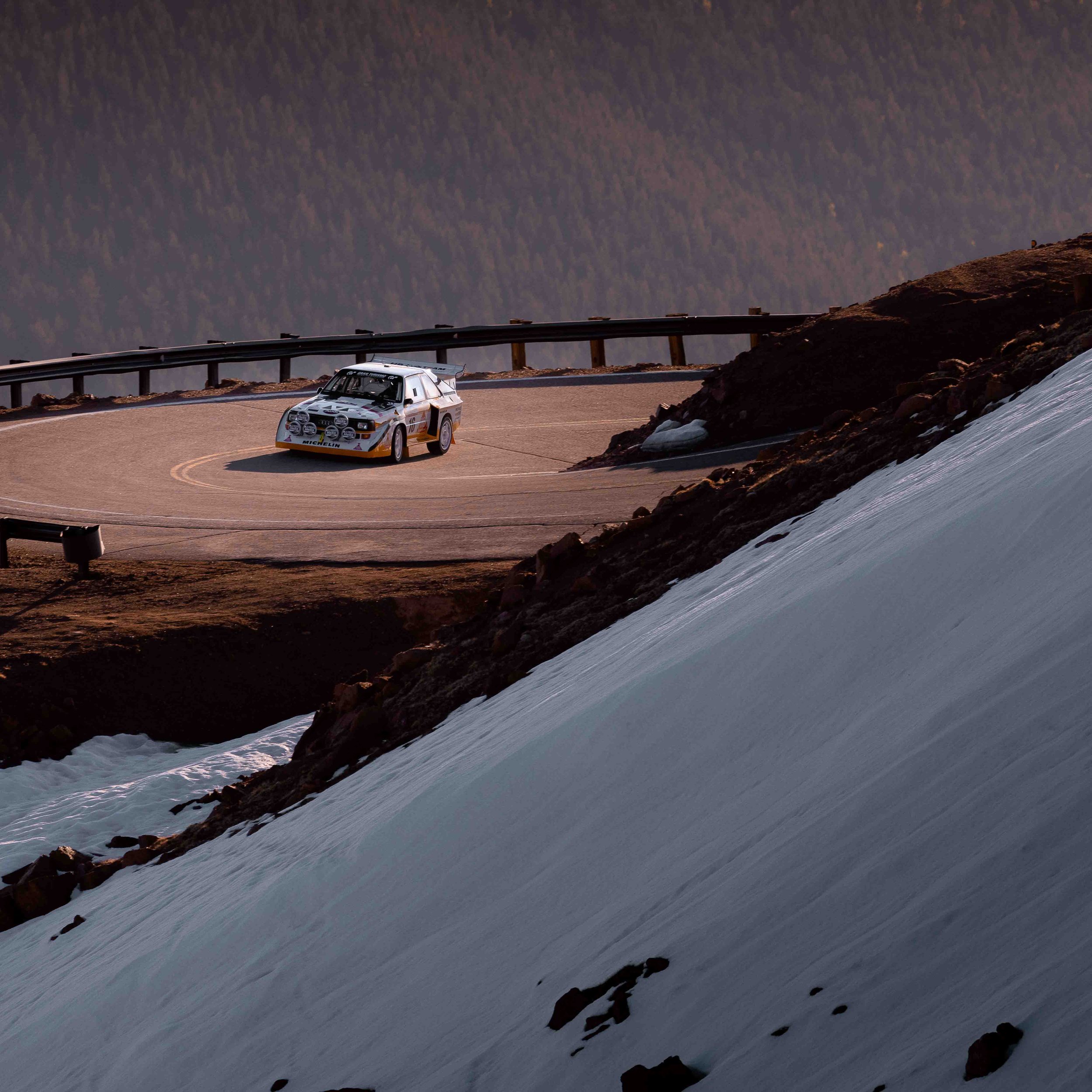 A rally car driving on a mountain road with a snow-covered slope on the right and a guardrail in the background.