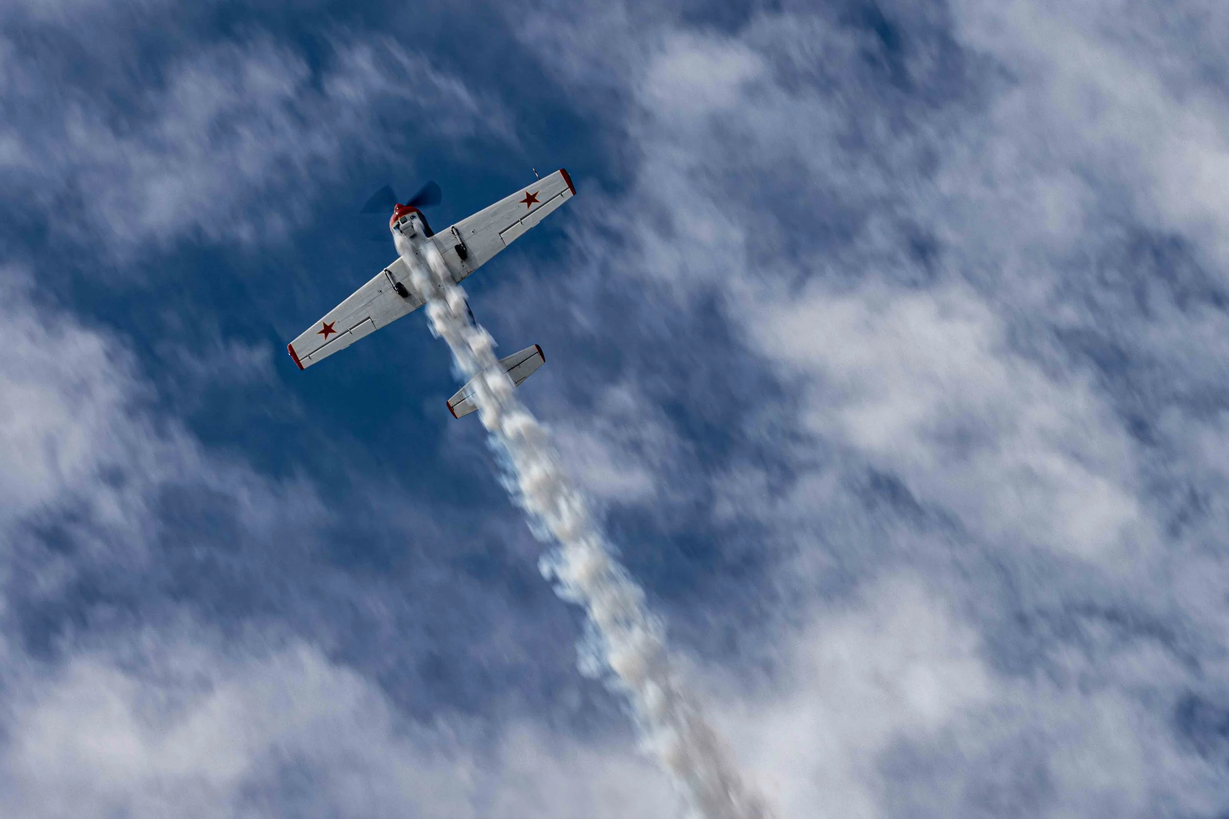 A vintage military airplane flying at high altitude, leaving white smoke trail behind against a sky with scattered clouds.