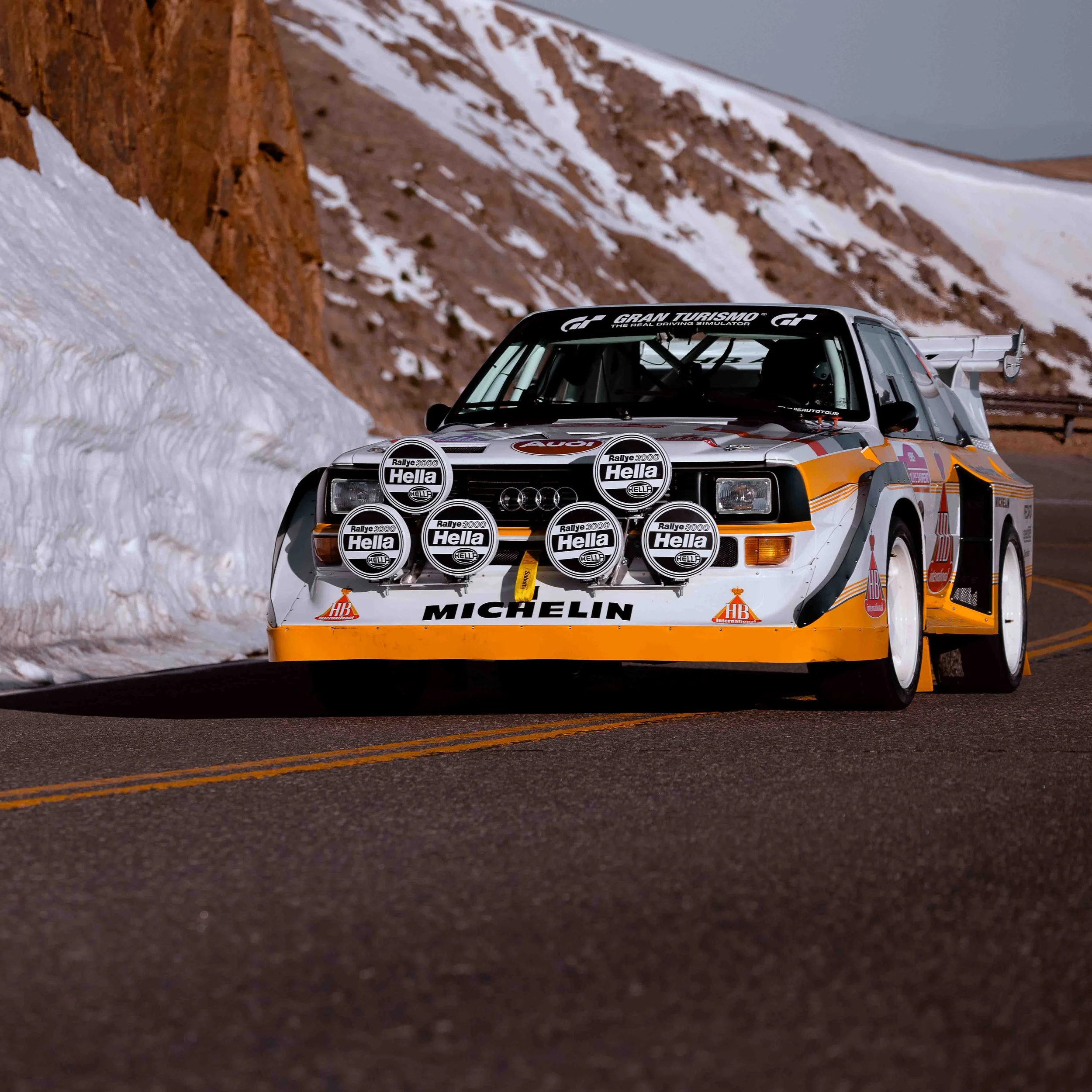 A vintage Audi rally car with a white and orange livery, equipped with multiple rally lights, racing on a mountain road with snow-covered mountain sides in the background.