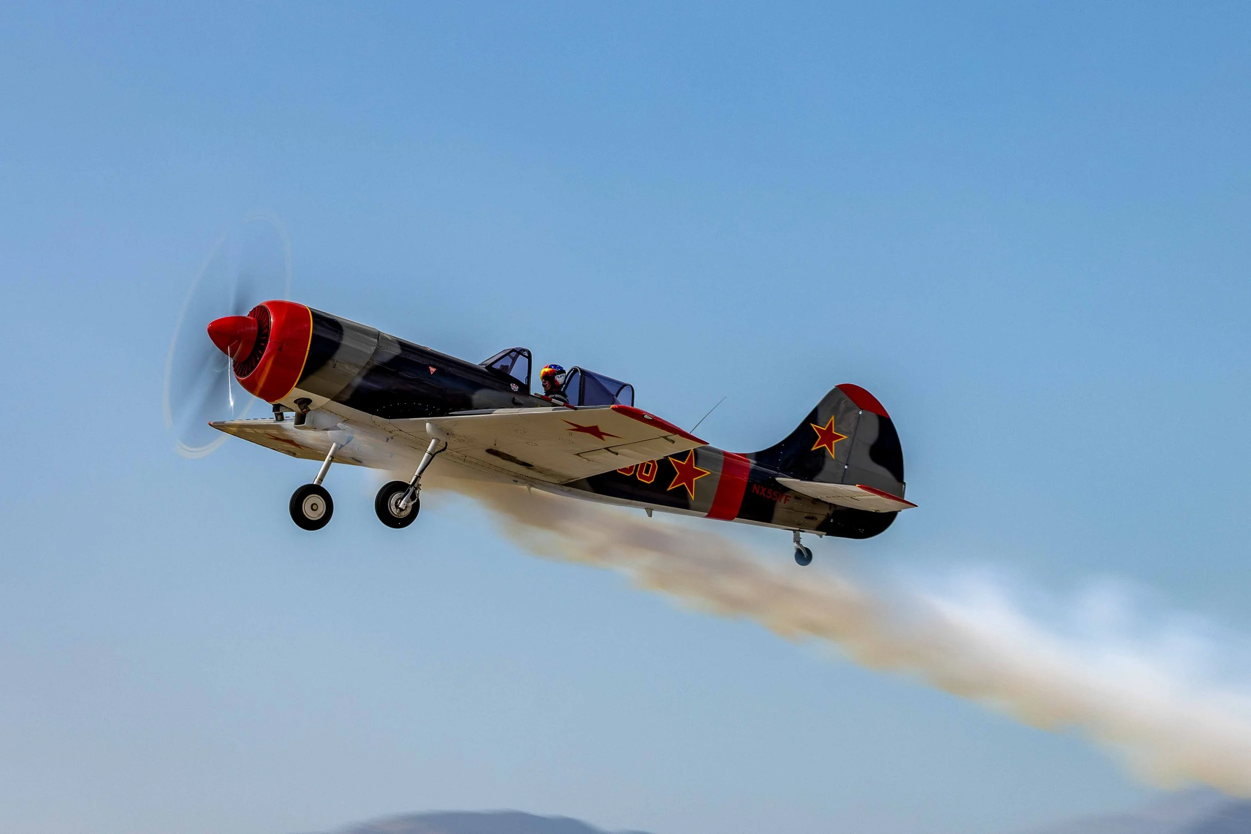 A vintage fighter jet aircraft flying in the sky, emitting white smoke trail, with a pilot visible in the cockpit.