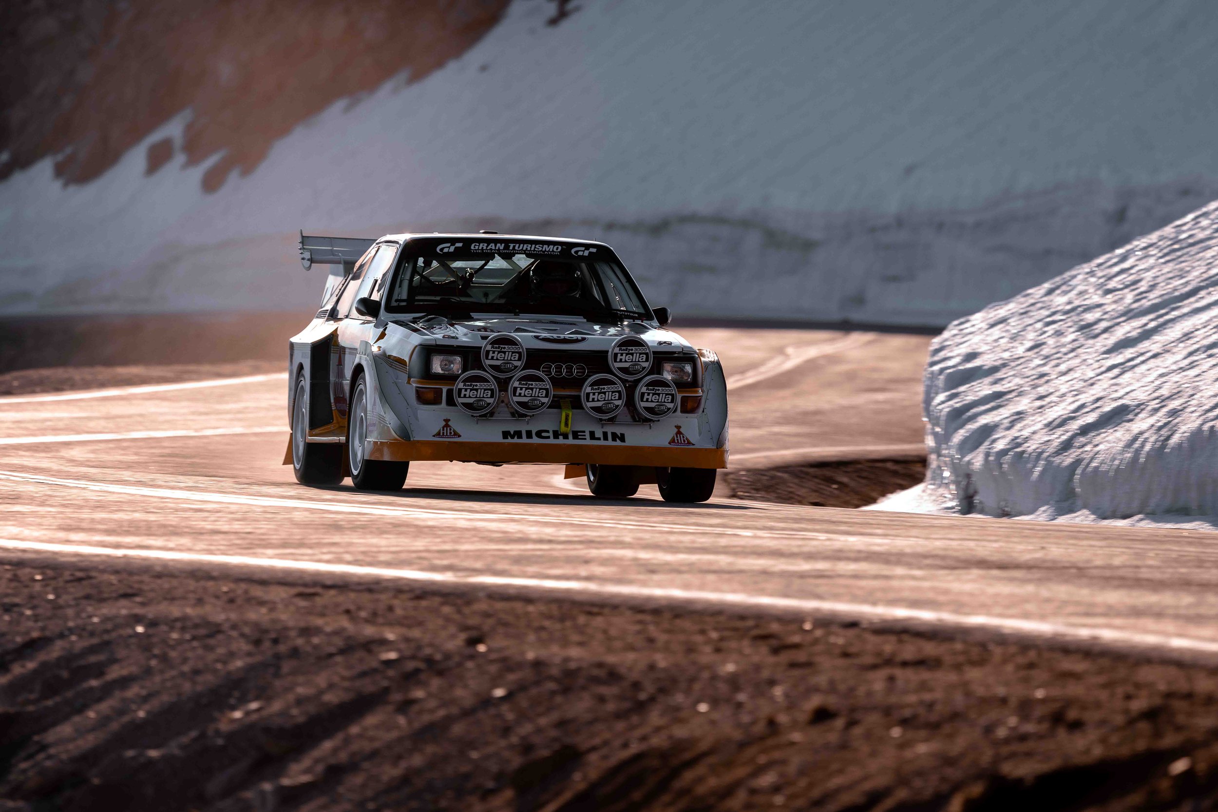 A vintage Audi rally car racing on a mountain road with snow on the sides, during daylight.