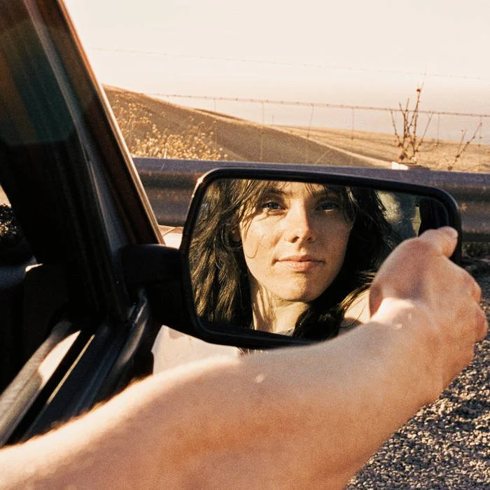 Person holding a side mirror of a car, looking at their reflection with a rural landscape in the background during sunset.