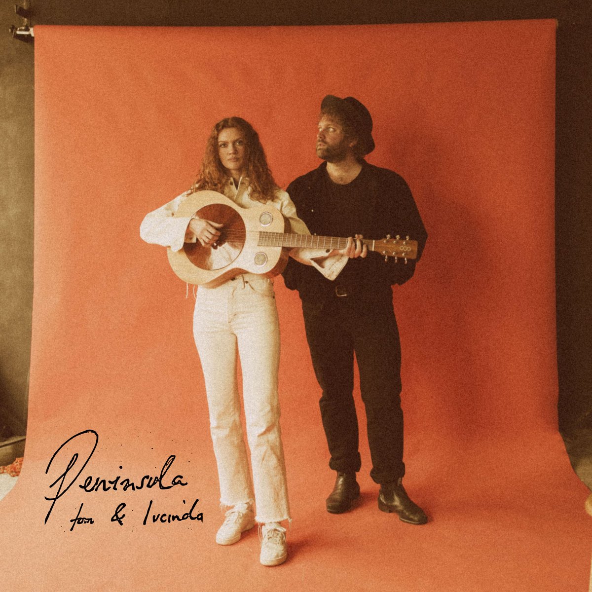 A woman and a man standing against an orange backdrop. The woman is holding a guitar and wearing white jeans and a white top, while the man is dressed in black and wearing a hat. The text 'Pentansula Tom & Lucinda' is handwritten on the bottom left corner.