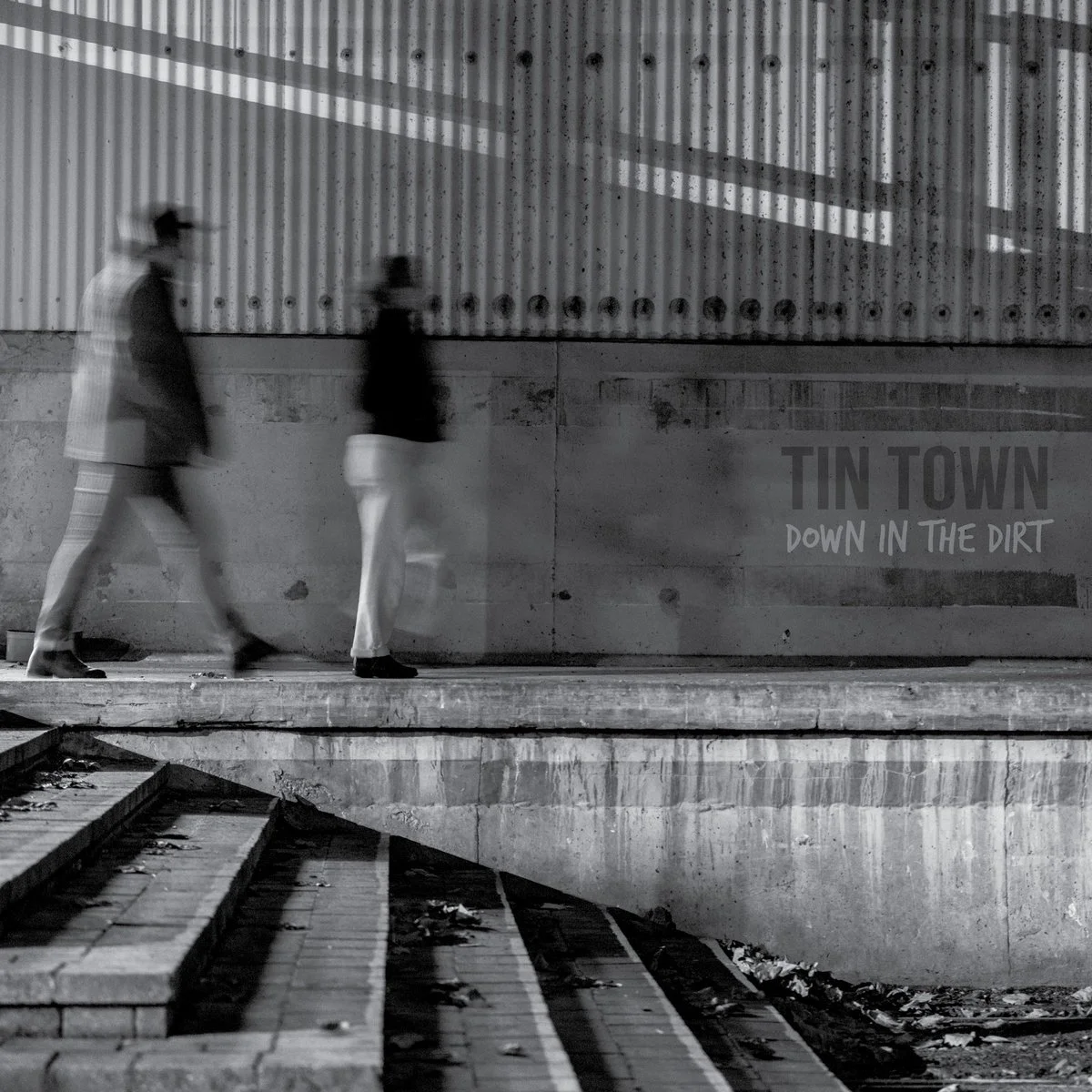 Two blurred people walking along a sidewalk in an urban setting, with a large wall in the background that has the words "TIN TOWN DOWN IN THE DIRT" written on it.