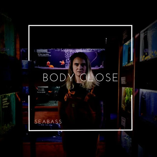 A woman standing in front of an aquarium display at a store, with the words 'BODY CLOSE' and 'SEABASS' overlaid.