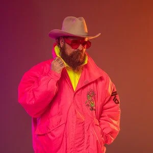 Person wearing a pink jacket, wide-brimmed hat, and red sunglasses, with a yellow shirt underneath, posing with hand near face against a gradient background.