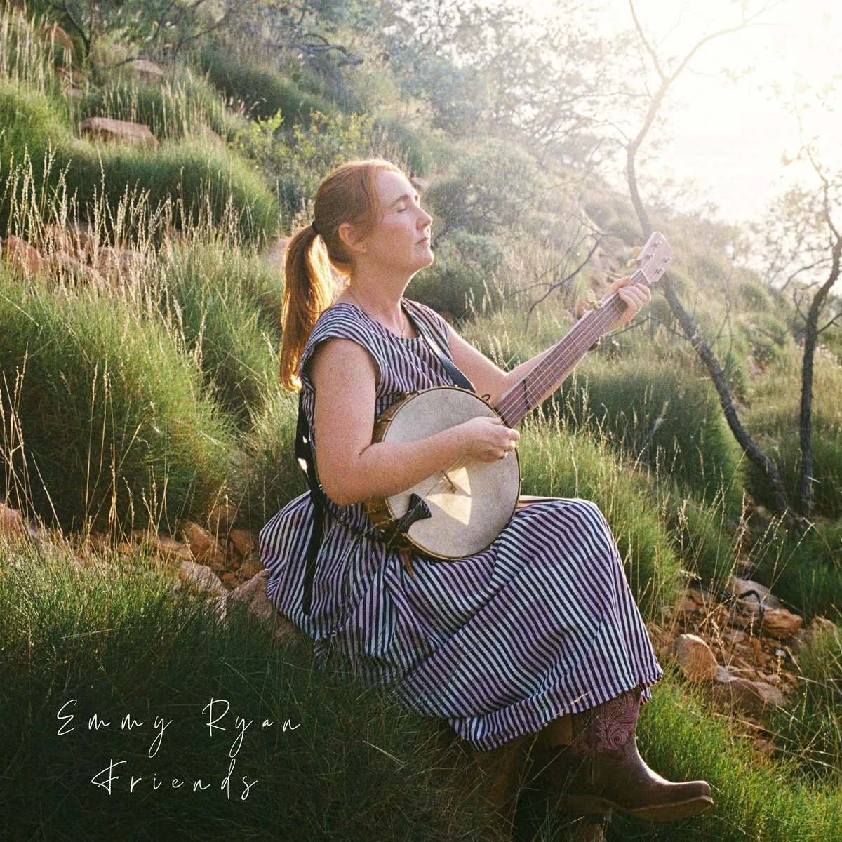 A woman with reddish hair tied back, dressed in a striped dress and cowboy boots, sitting on a grassy hill playing a banjo during sunset in a natural outdoor setting.