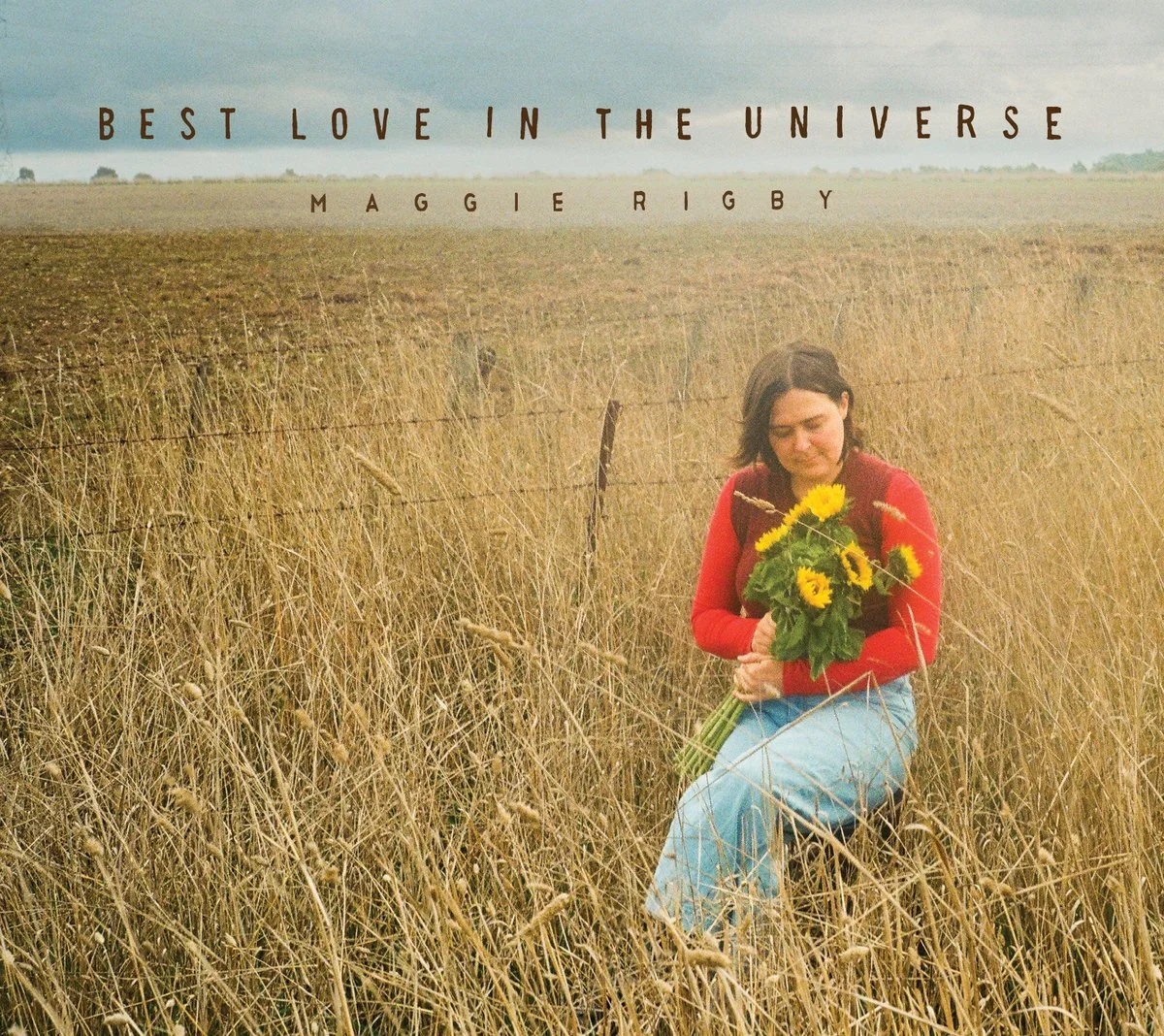 A woman kneeling in a field of tall, dry grass holding a bouquet of bright yellow sunflowers, with a cloudy sky and distant horizon in the background.