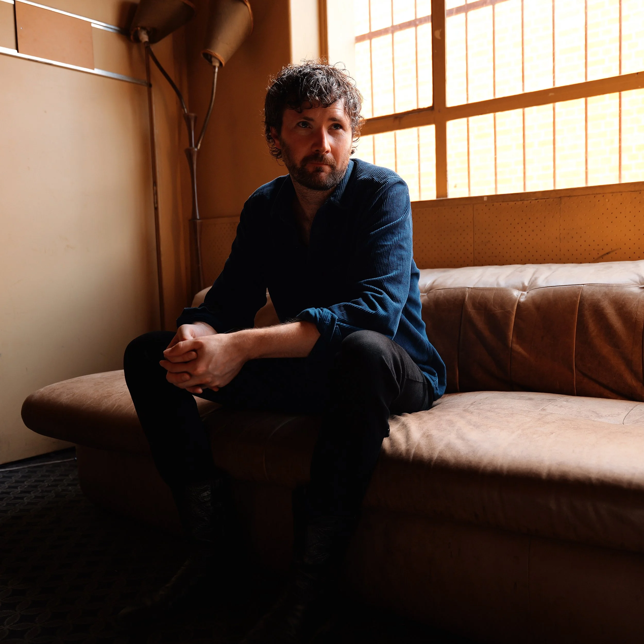 A man sitting on a brown leather couch with natural light streaming through a large window with wooden grid patterns in the background.
