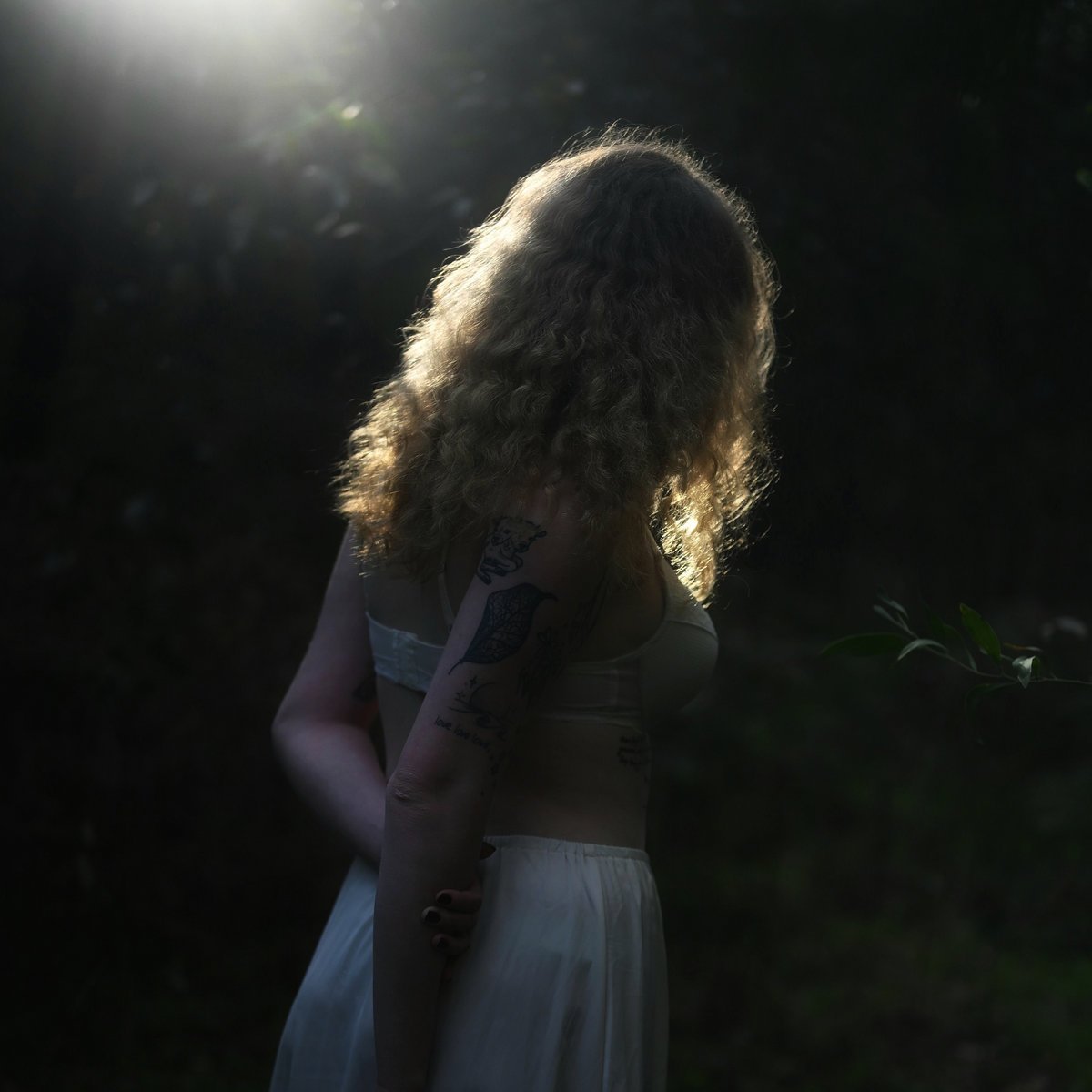 A woman with curly blonde hair, tattoos on her arm, wearing a white dress, standing outdoors in a dark forest with sunlight shining behind her.