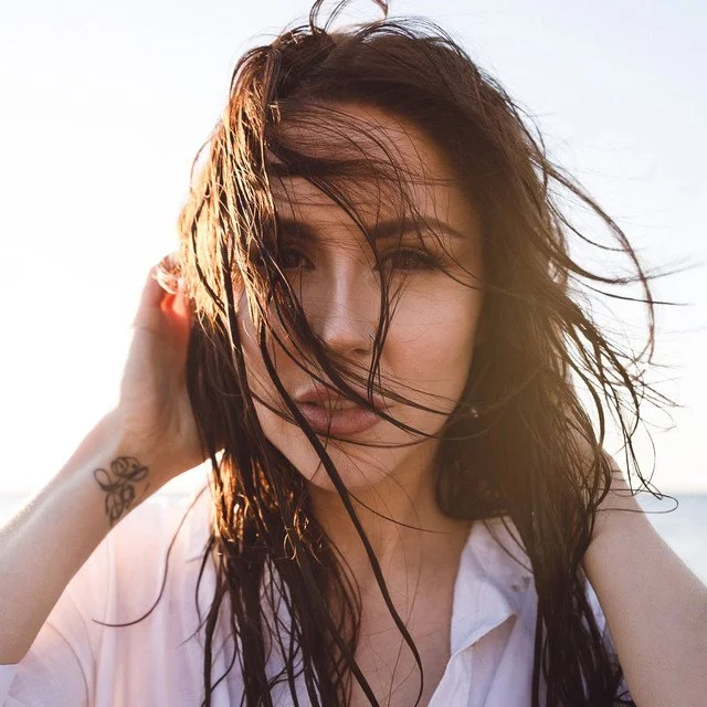 A woman with wet hair and a white shirt, posing outdoors with wind blowing her hair.