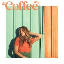 A woman with long hair wearing an orange top and green pants, standing against a colorful striped wall.