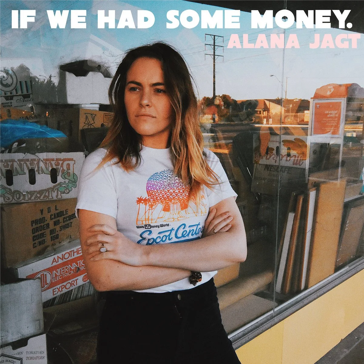 A woman with shoulder-length hair and crossed arms standing outside a shop, wearing a souvenir T-shirt featuring Disneyland's Epcot Center, with boxes and storefront reflected in the window behind her.