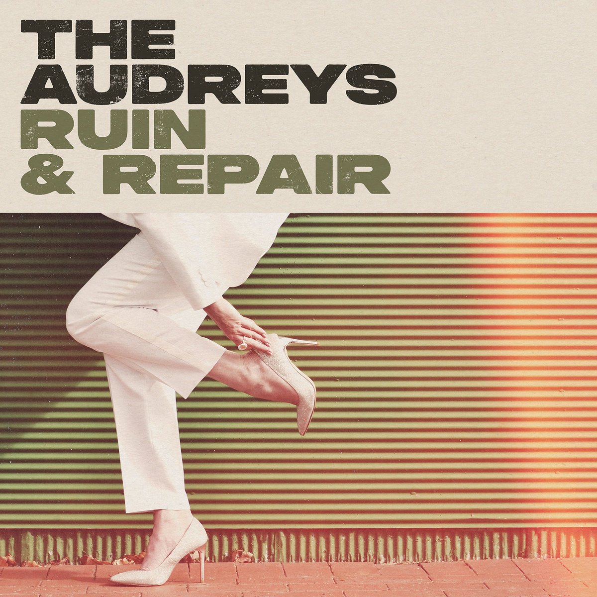 Text overlay reading 'The Audreys Ruin & Repair' over a photo of a person in a white suit and high heels adjusting their shoe against a green corrugated wall.