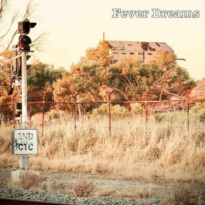A railway crossing signal with lights and a warning sign, an overgrown field with dry grass, a line of trees, and a partially visible abandoned building in the background. The words "Fever Dreams" are written in the top right corner.
