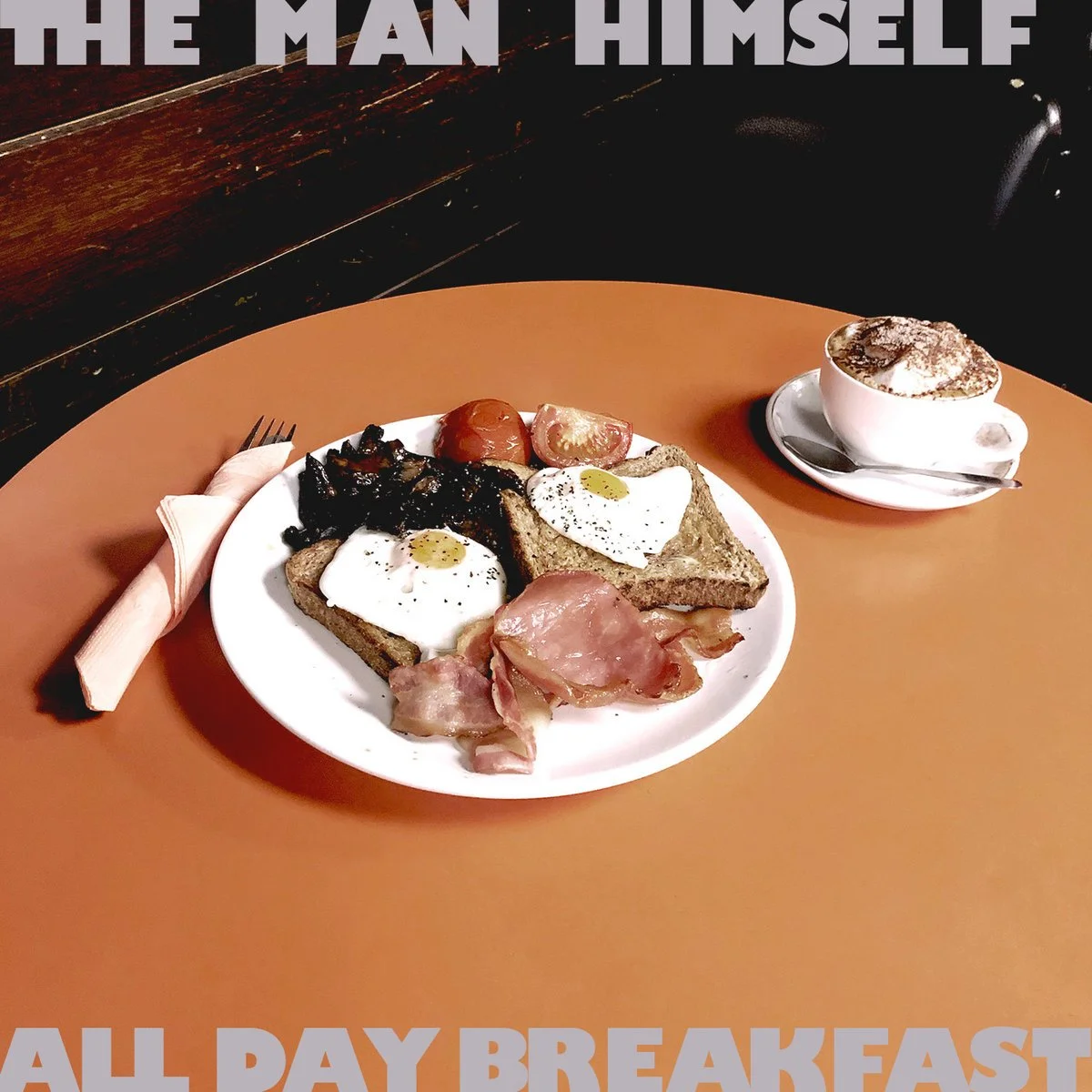 A breakfast plate with fried eggs, bacon, toast with eggs, tomatoes, and sautéed mushrooms, accompanied by a cup of coffee with frothy foam.