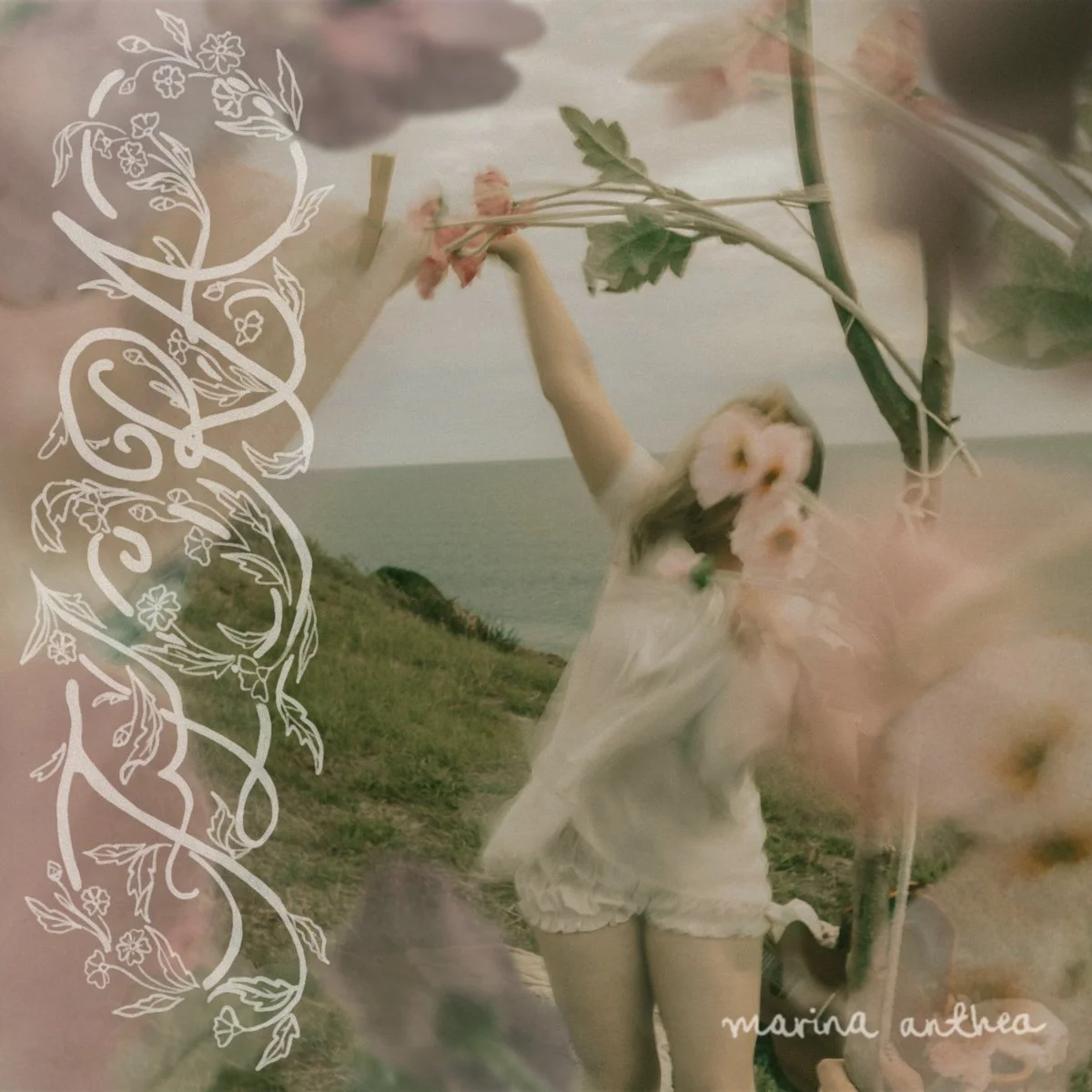 A girl holding flowers stands outdoors near the coast, with a grassy hill and the ocean in the background. The photo has an artistic overlay of white floral and vine designs, and the text 'marina anthea' in the lower right corner.