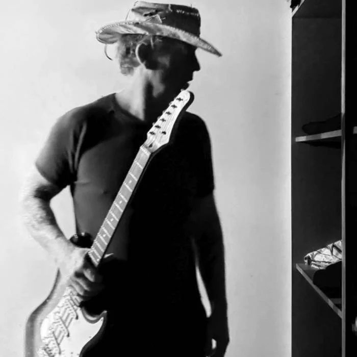 A person wearing a straw hat and glasses holding an electric guitar, standing indoors near shelves or a closet.