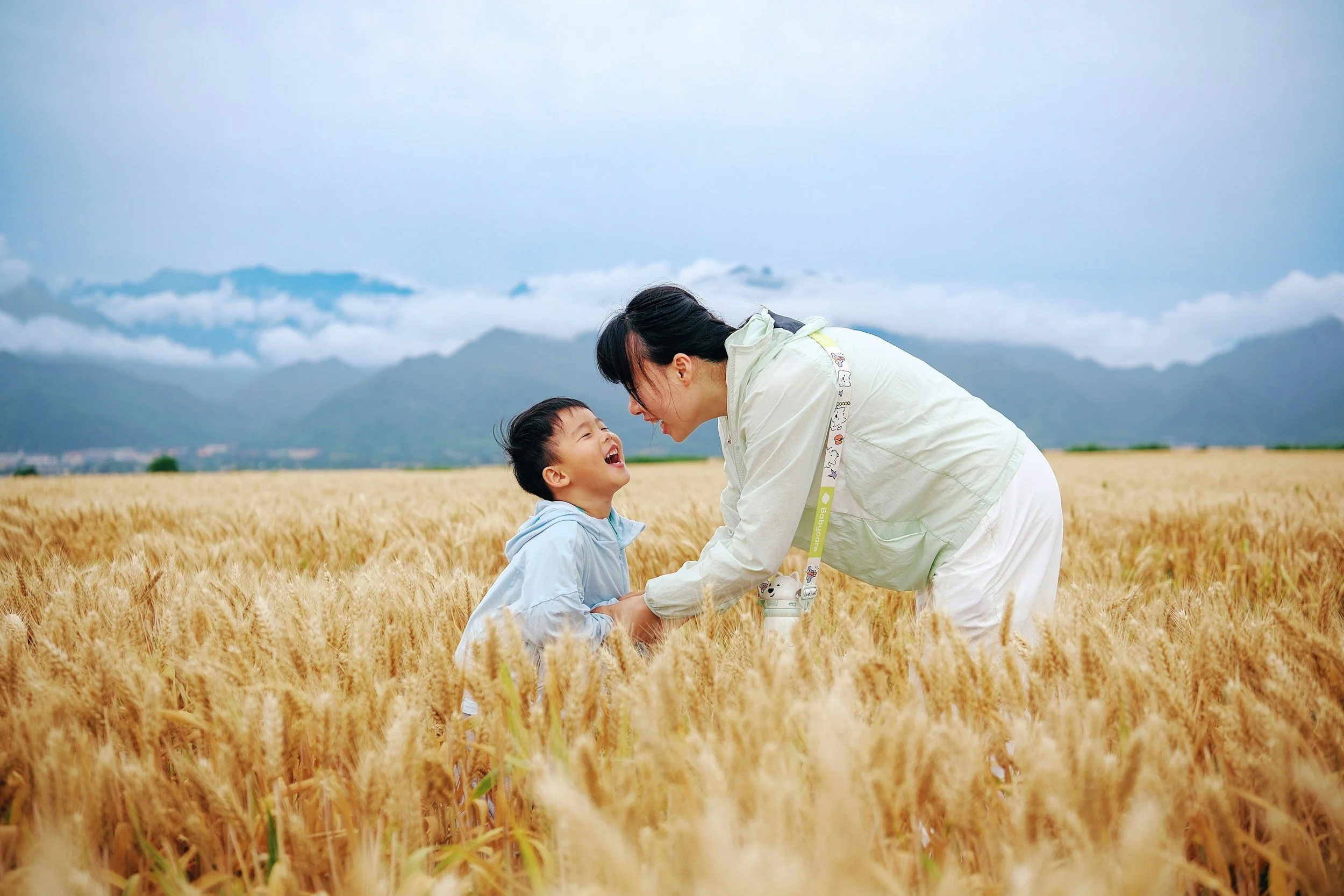 A woman and a young boy are smiling and laughing together in a wheat field with mountains and cloudy sky in the background.