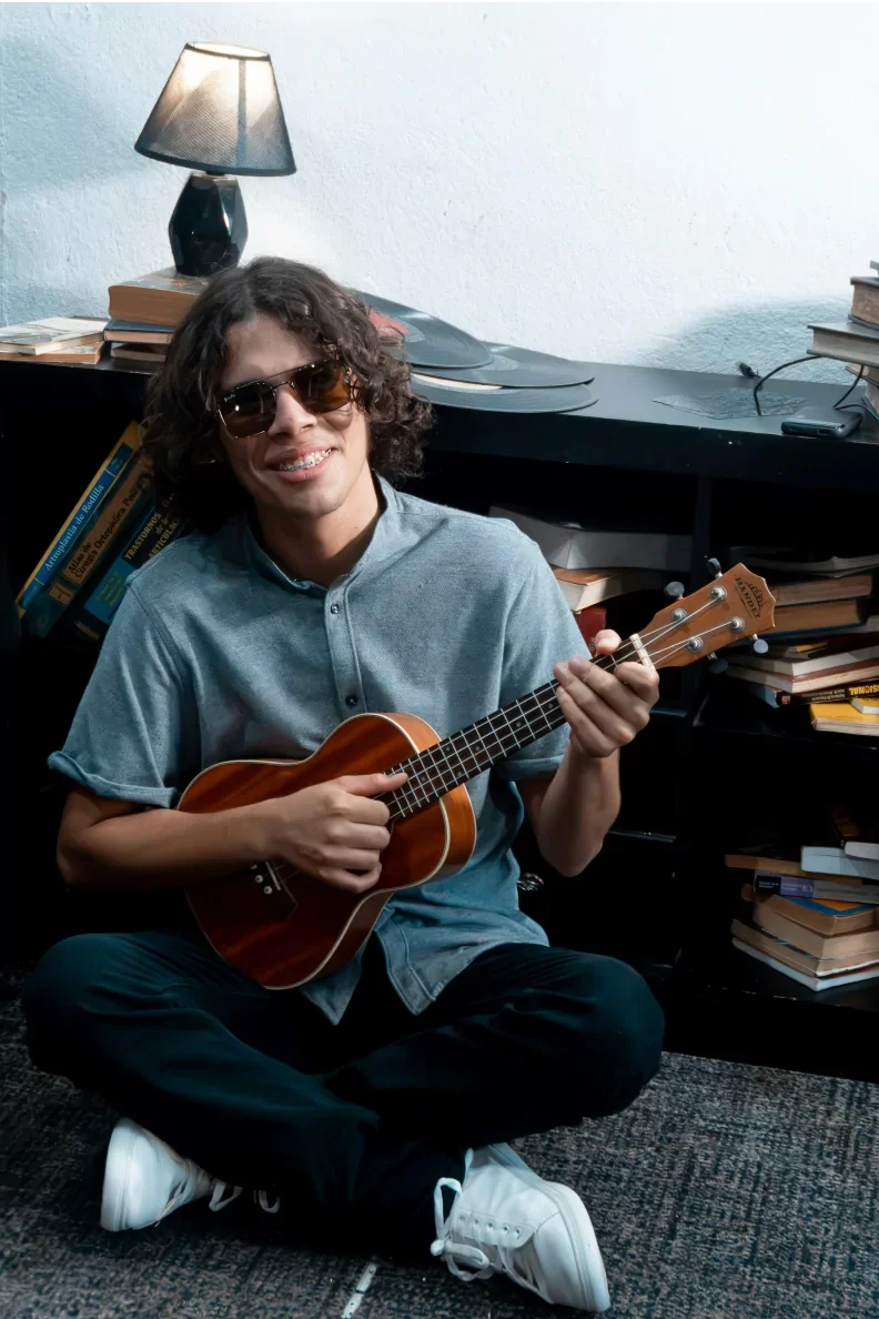 A young man with curly hair, wearing sunglasses and braces, sitting cross-legged on the floor, holding a small acoustic guitar, in a room with a bookshelf, books, and a desk lamp.