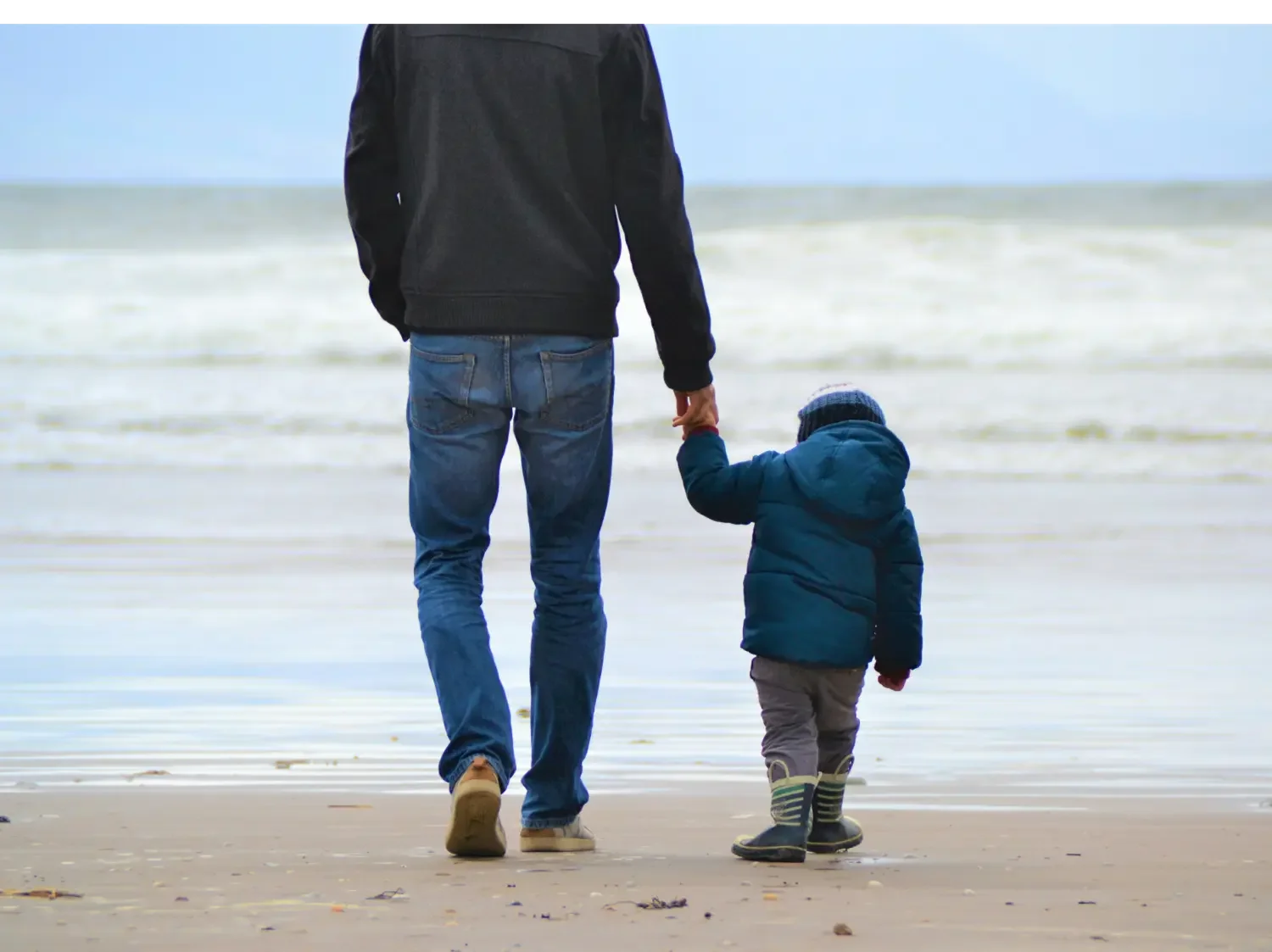 Rear view of a parent and young child holding hands while walking together on a sandy beach toward the ocean waves.