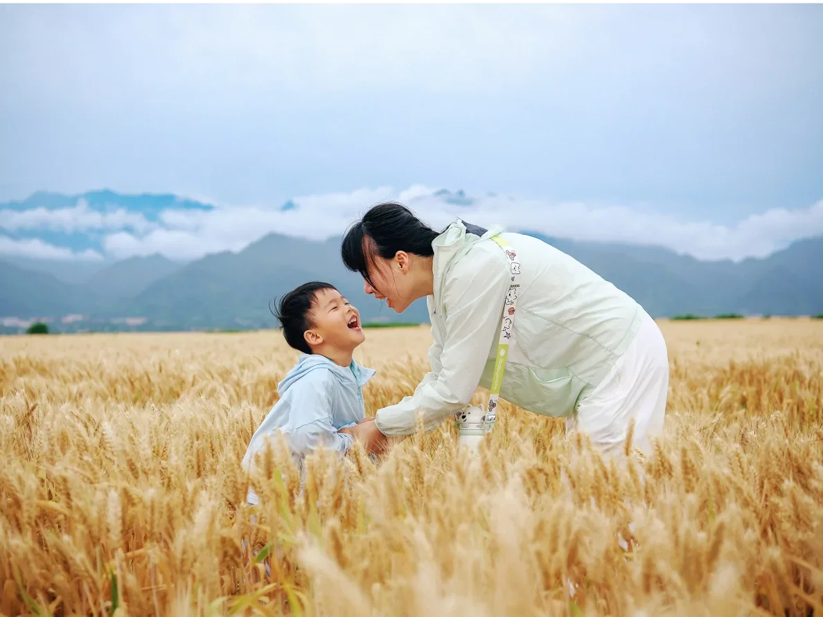 A woman and a young boy are smiling and laughing together in a wheat field with mountains and cloudy sky in the background.